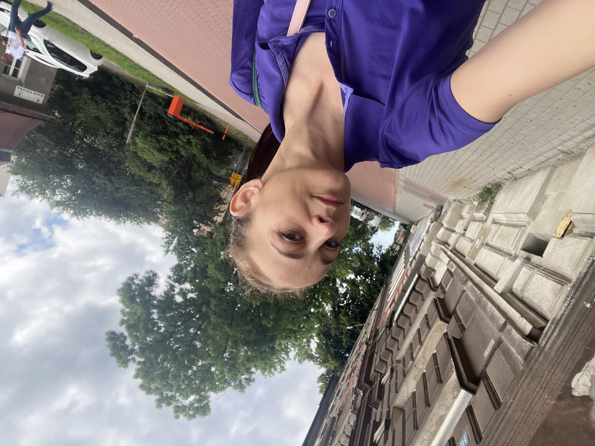 A woman is captured in a selfie, taken from a low angle, on a sidewalk in a city. She has light skin and brown hair pulled back. She is wearing a purple polo shirt. The background shows a building with ornate detailing on its facade, a tree-lined street, and a cloudy sky. A red barrier and a white car are visible further down the street.