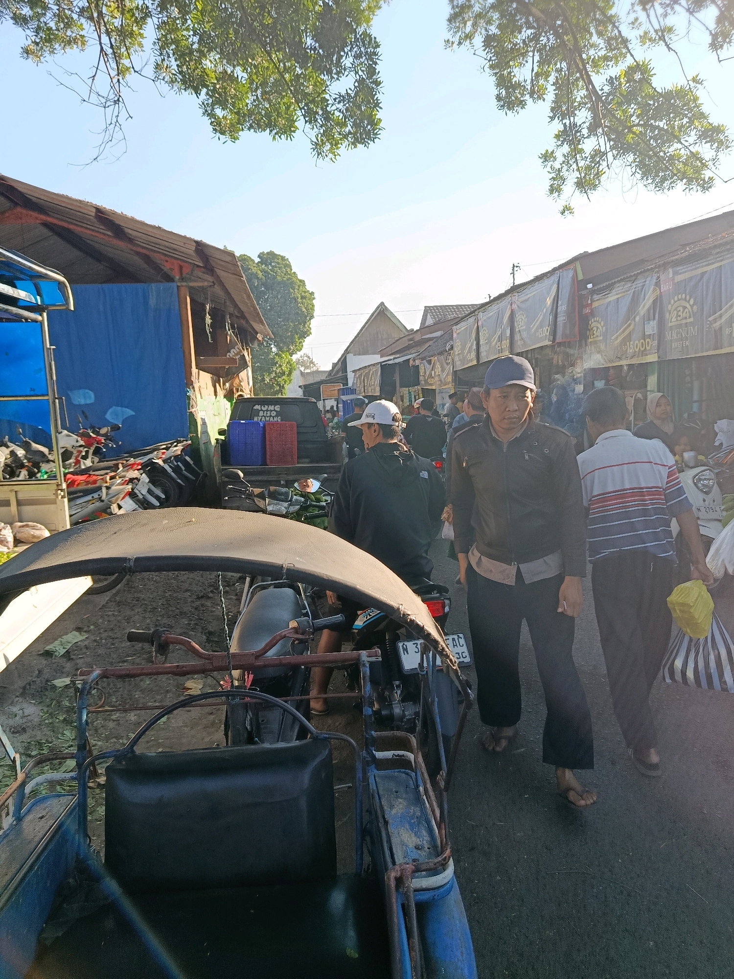 In Bululawang, Indonesia, a street market scene unfolds under a bright, sunny sky with dappled sunlight filtering through overhead foliage. The foreground is dominated by a blue and black motorized tricycle, its canvas roof casting a shadow. To its right, a man wearing a black jacket and a blue cap stands looking forward, while another man in a striped shirt walks away from the camera. Behind them, a black pickup truck is parked, its bed loaded with blue and red plastic crates. A row of market stalls with banners advertising "234 Magnum Kretek" lines the background, with various people browsing and interacting. The ground is a mixture of asphalt and dirt, with scattered leaves and debris. Several motorcycles are parked along the street, suggesting it is a busy market day. The overall impression is a lively, everyday scene in a local Indonesian market.