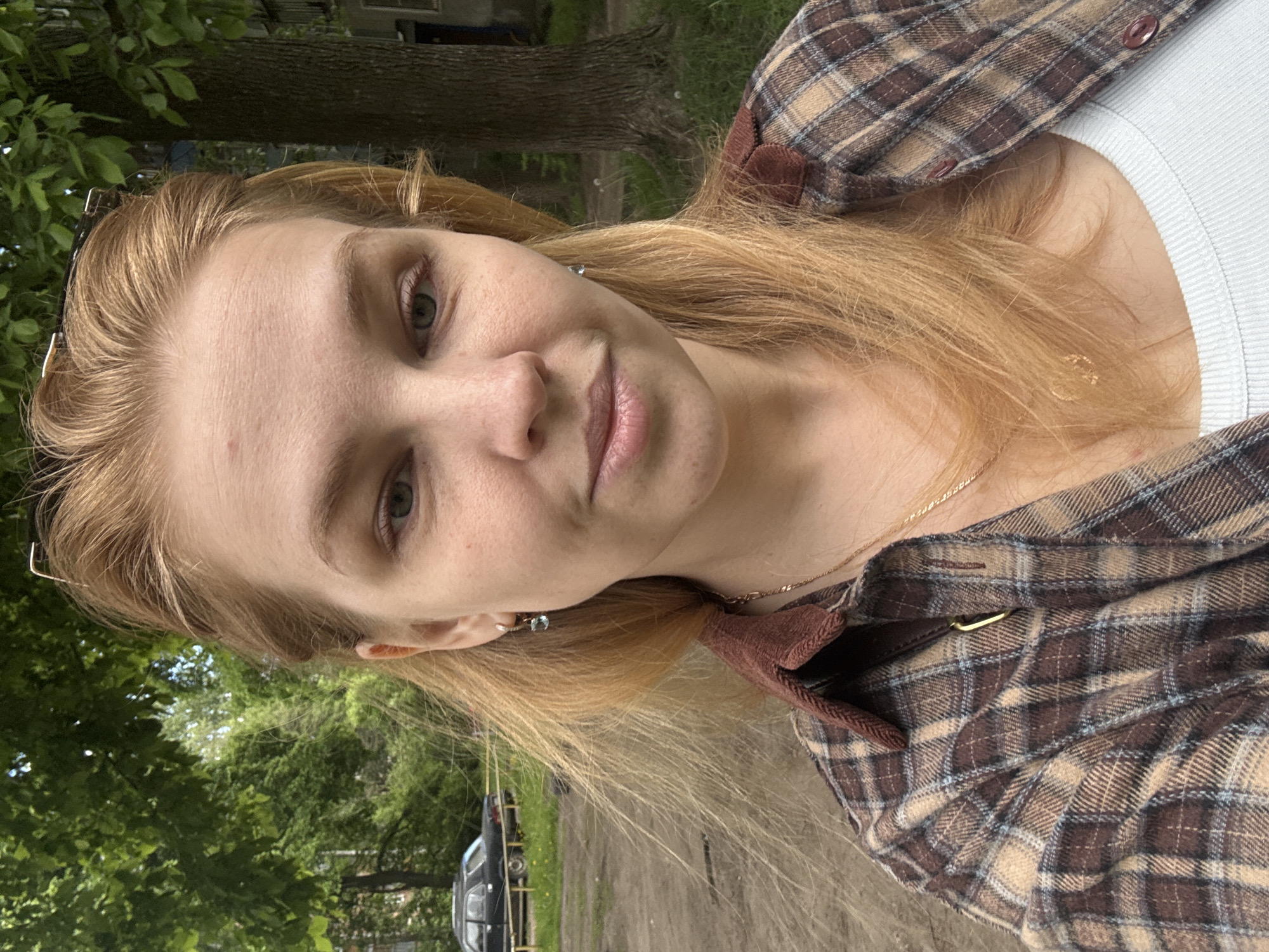 A young woman with light brown to blonde hair and light-colored eyes is captured in a close-up, outdoor selfie. She looks directly at the camera with a subtle, gentle smile. She is wearing a brown and tan plaid flannel shirt, with a light-colored, possibly white or light blue, ribbed top visible underneath. A thin gold-colored necklace adorns her neck, and a small earring is visible in her left ear. Her hair is worn down.
The setting appears to be outdoors during daytime, with ample green foliage and trees in the background, suggesting a natural or park-like environment. In the mid-ground, a dirt or unpaved path is visible, and a dark-colored vehicle, possibly an older car or van, is parked behind a low yellow barrier. The overall impression is casual and relaxed. Given the provided context, the scene is located in Rusanovo, Russia. The light is soft, indicating it might be an overcast day or late afternoon. No visible text is present in the image.