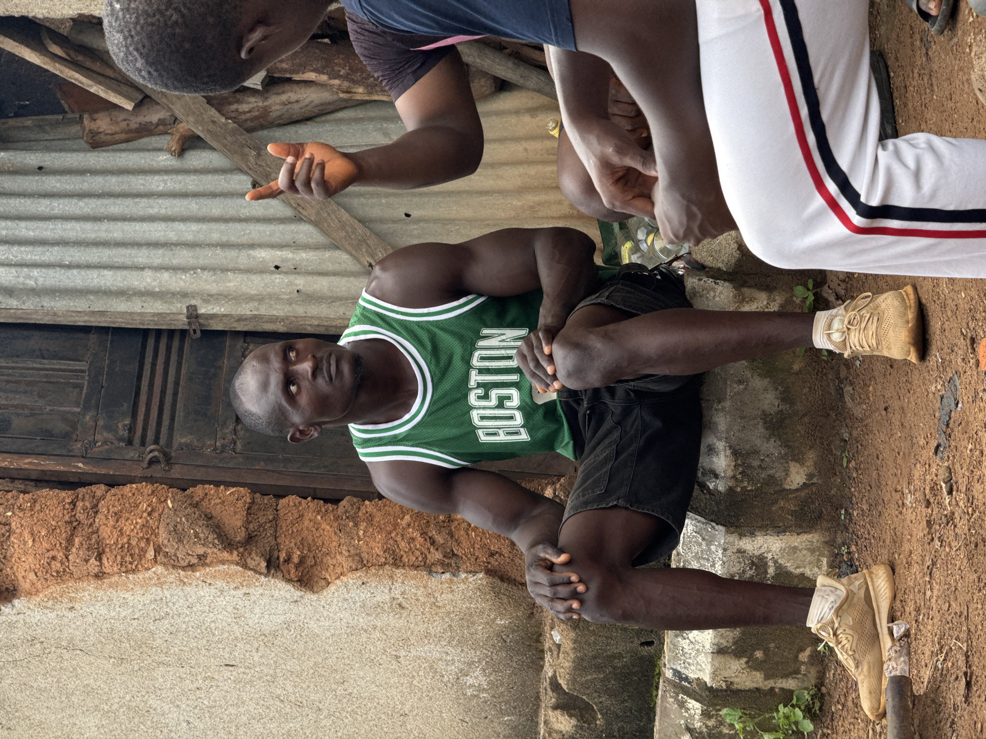 In Akwanga, Nigeria, two men are seated outdoors, engaged in conversation. The man on the left is wearing a green "Boston" basketball jersey and black shorts, with tan athletic shoes. He is sitting on a concrete step. The man on the right is wearing a dark shirt and white pants with red and black stripes. His left hand is raised, index finger pointing, as he speaks. The background consists of a textured wall made of reddish-brown mud and lighter plaster, a dark metal door, and corrugated metal siding with wooden planks. The ground is made of dirt and gravel. Several plastic bottles are visible on the ground near the seated men.