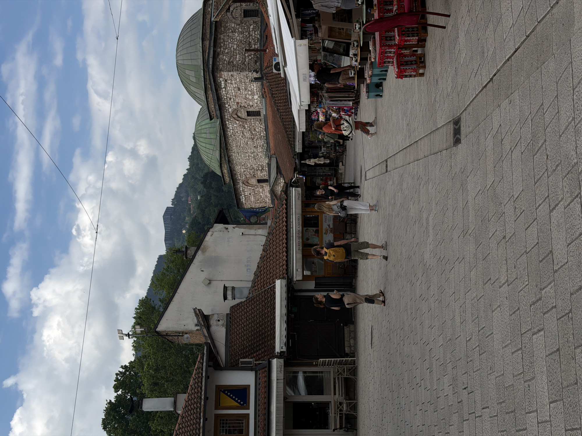 The image depicts a view of Baščaršija, the historic bazaar in Sarajevo, Bosnia and Herzegovina. In the foreground, the cobblestone pavement is visible, with a slight depression indicating a drainage channel. Several people are walking across the square. On the left, a building with a tiled roof and windows displays the Bosnian flag. To the right of this building, a food establishment with a "Coca-Cola" advertisement is visible. Further to the right, market stalls with awnings are set up, displaying various goods. In the background, the iconic green domes of the Gazi Husrev-beg Mosque complex are prominent, constructed from stone. The sky is partly cloudy, with white cumulus clouds against a blue backdrop. Trees and a hillside with buildings are visible in the distance. The overall scene captures a bustling atmosphere in a historical urban setting.