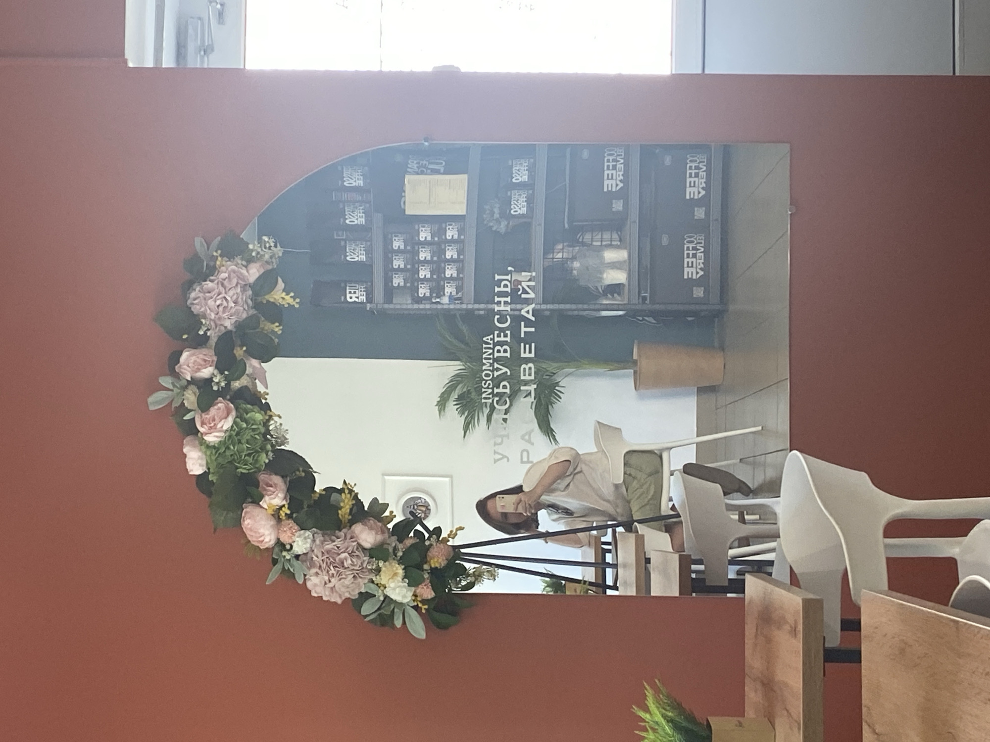 The image shows an interior scene, likely a cafe or restaurant, in the city of Chaikovsky, Russia, as evidenced by the Cyrillic text in the image. The focal point is an arched mirror adorned with a floral wreath of pink and white flowers. The reflection in the mirror reveals a partial view of the interior, including shelving stocked with "COFFEE" boxes, interior decoration and plants with Cyrillic signs "INSOMNIA" and more Cyrillic text which may be a slogan, and a person taking a selfie with a phone. The mirror's reflection also shows several white chairs and wooden tables. The wall behind the mirror is painted a terracotta color. A fragment of a wooden structure or furniture is visible in the foreground.