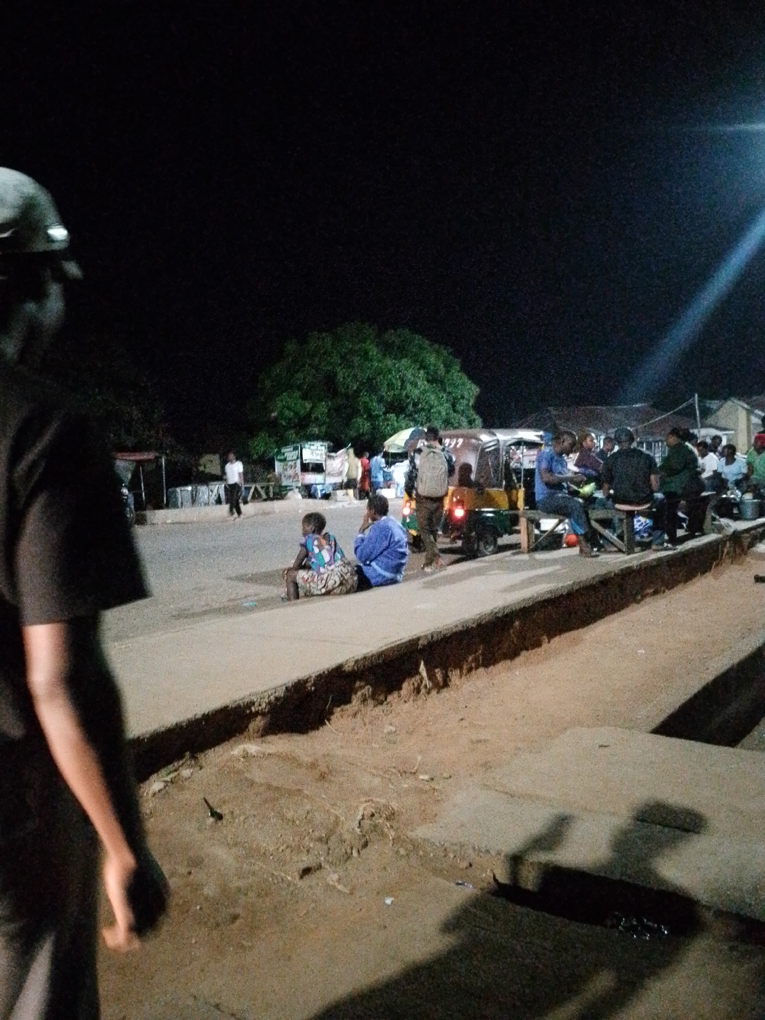 The image depicts a nighttime scene on a street in an unknown city. In the foreground, a person wearing a cap and a dark shirt is partially visible from the side, with their back to the camera. The mid-ground shows a street with several people gathered. Some individuals are seated on what appear to be benches or low walls, while others are standing. A three-wheeled vehicle, possibly a tuk-tuk, is parked on the street, with people around it. There are stalls or small shops lining the street, indicated by structures and some illuminated signage in the background. A large tree is visible behind some of the stalls. The ground appears to be paved with concrete and dirt. The lighting suggests artificial illumination from streetlights or nearby sources.