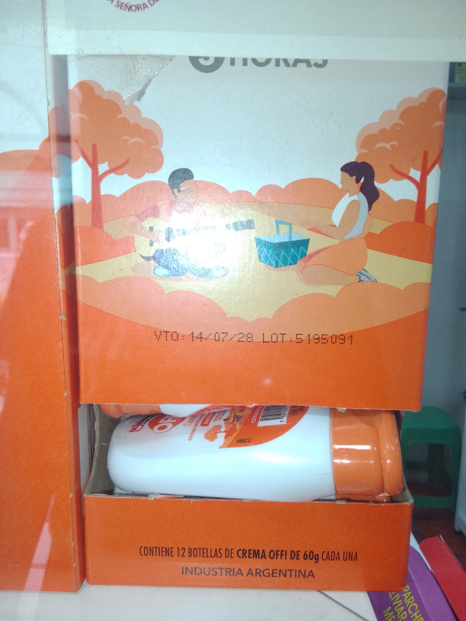 This image shows a close-up of an orange cardboard box containing bottles of a product labeled "CREMA OFF!". The box has an illustration of a picnic scene with stylized trees and figures. Text on the box indicates "VTO: 14/07/28 LOT: 5195091" and "CONTIENE 12 BOTELLAS DE CREMA OFF! DE 60g CADA UNA INDUSTRIA ARGENTINA". The product bottles are white with orange caps. The image is taken indoors, with a green stool visible in the background on the right. There is no information in the image that indicates the location of Juan Griego, Venezuela.