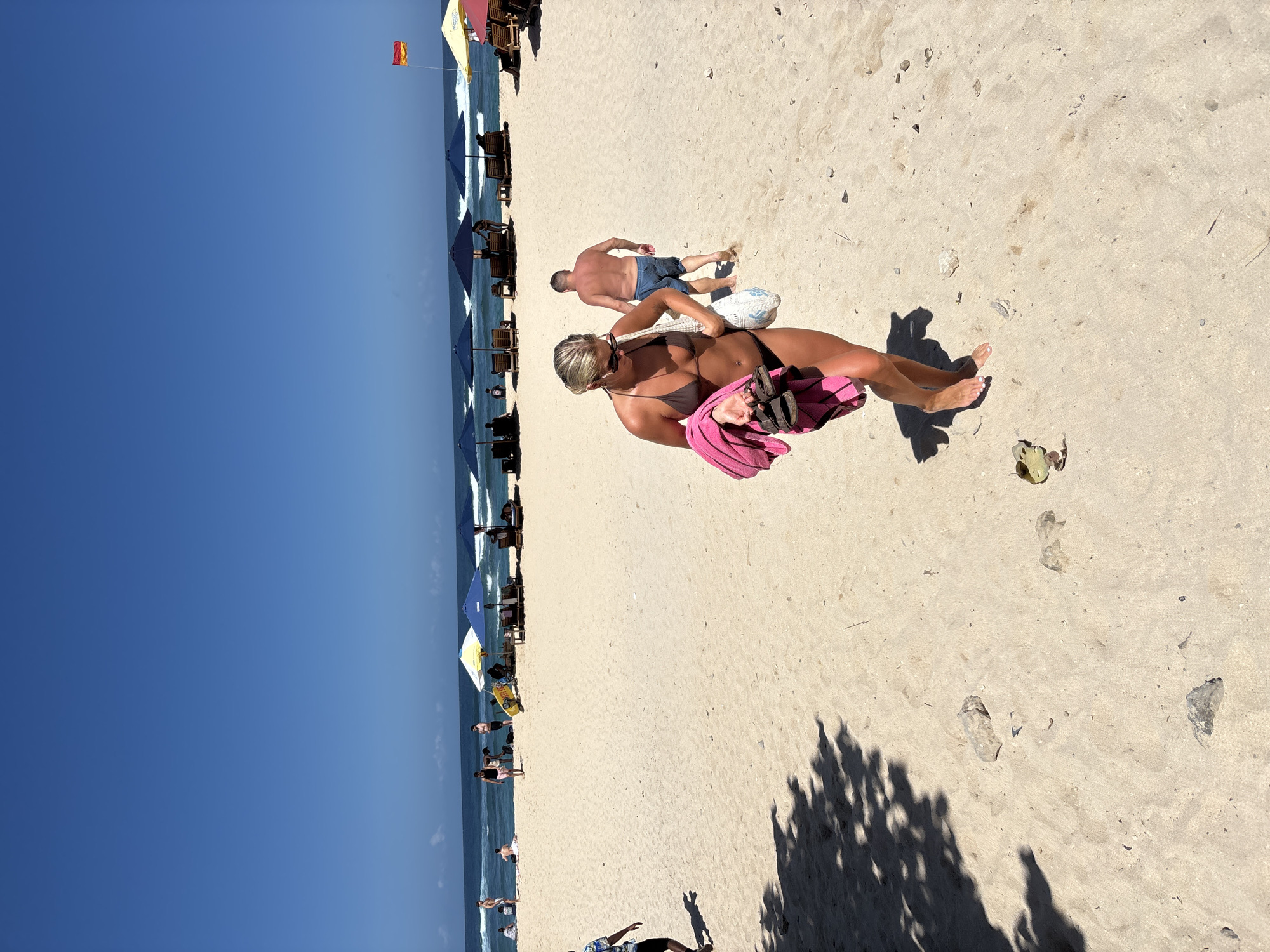 In Kuta Selatan, Indonesia, a bright, sunny day at the beach unfolds. The sand is light and dry, with scattered small stones and debris. In the foreground, a woman with blonde hair, wearing sunglasses and a brown bikini, walks towards the camera. She is carrying a pink towel draped over her left arm and a pair of dark sandals in her hand. Behind her, a man in denim shorts and a bare chest walks away from the camera.
Further back, several beach umbrellas, mostly blue and some yellow, are set up on the sand. Some beach chairs are visible under the umbrellas. People are scattered across the beach, some sitting or standing near the umbrellas, and others are visible in the distance by the ocean. The ocean is a deep blue with gentle waves rolling onto the shore. The sky is a clear, vibrant blue. A red and yellow flag is flying from a pole on the right side of the image, likely indicating a lifeguard station or hazard warning. The overall scene depicts a typical day at a popular beach resort.