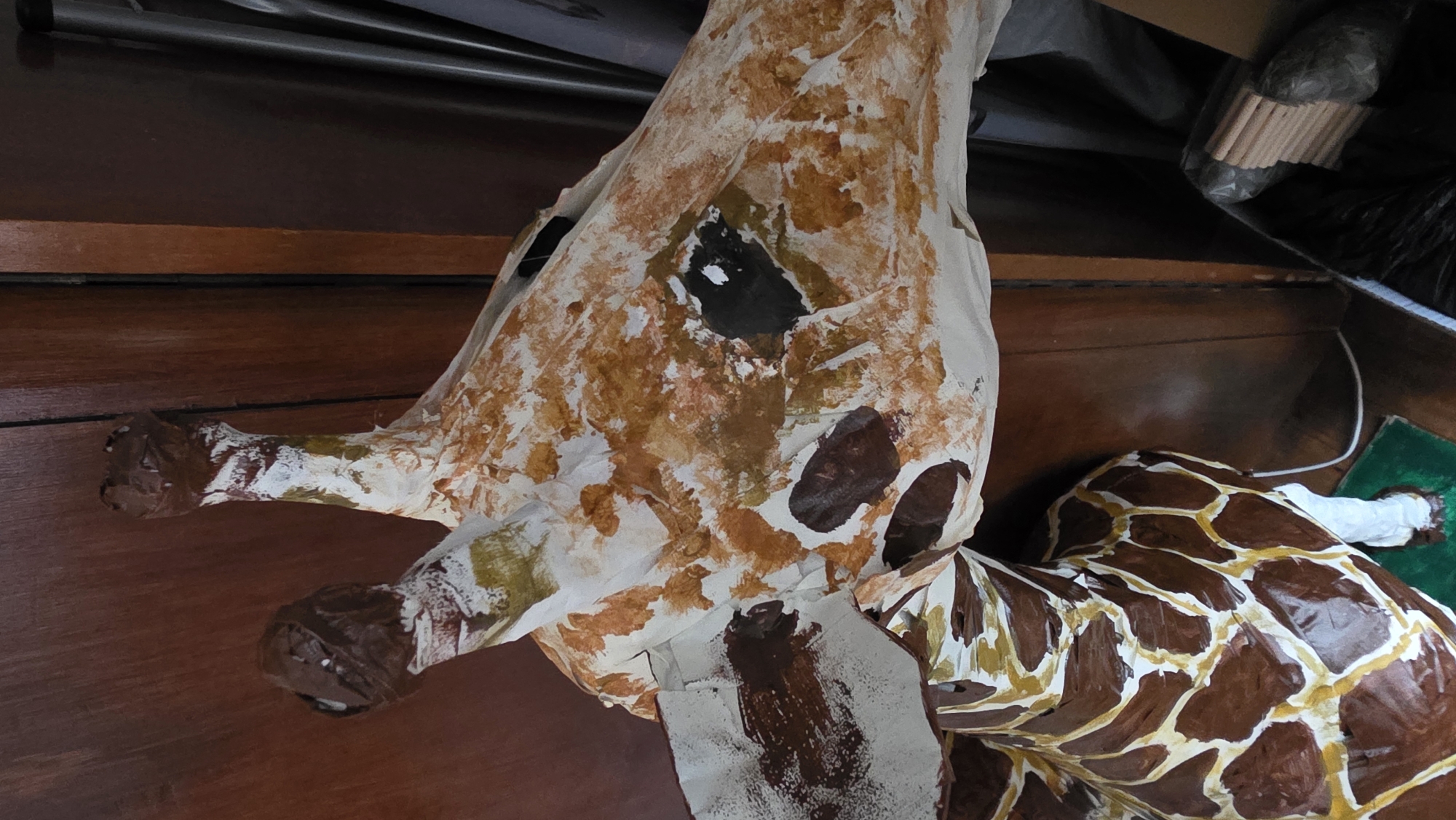 The image shows a partially completed papier-mâché giraffe head and torso resting on a dark brown wooden surface. The head is painted with a pattern of light brown and cream spots, and it has a large, irregular black hole near what would be the eye area. The horns are painted brown and the surface shows areas where the paint application seems unfinished. The torso is painted with a classic giraffe pattern of dark brown spots outlined in cream. Other objects in the background include a bundle of wooden dowels wrapped in clear plastic, a green object, a white cord, and other unidentified materials suggesting a storage area or workshop. There is no information to indicate that this image was taken in Blagoevgrad, Bulgaria.