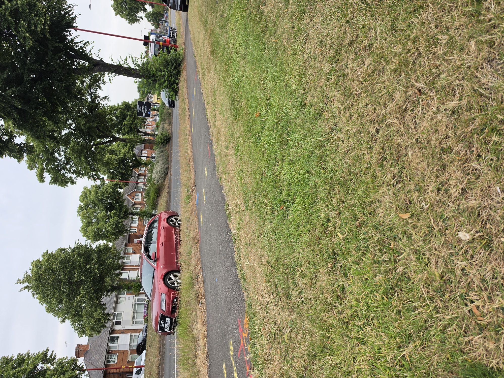 A red Audi A4 Avant estate car is parked on the side of a road in Birmingham, United Kingdom. The car is a right-hand drive vehicle with the license plate "RV61 ZBZ". The car is positioned on the grass verge, facing slightly to the left. A paved path runs parallel to the road, with yellow and red painted markings on it.
In the background, there is a row of residential houses with red brick walls and tiled roofs. Large green trees are visible on either side of the road, and the sky is overcast. A signpost with directional arrows is also visible. Further down the road, a truck and other vehicles can be seen. The scene depicts a typical suburban street on a cloudy day.