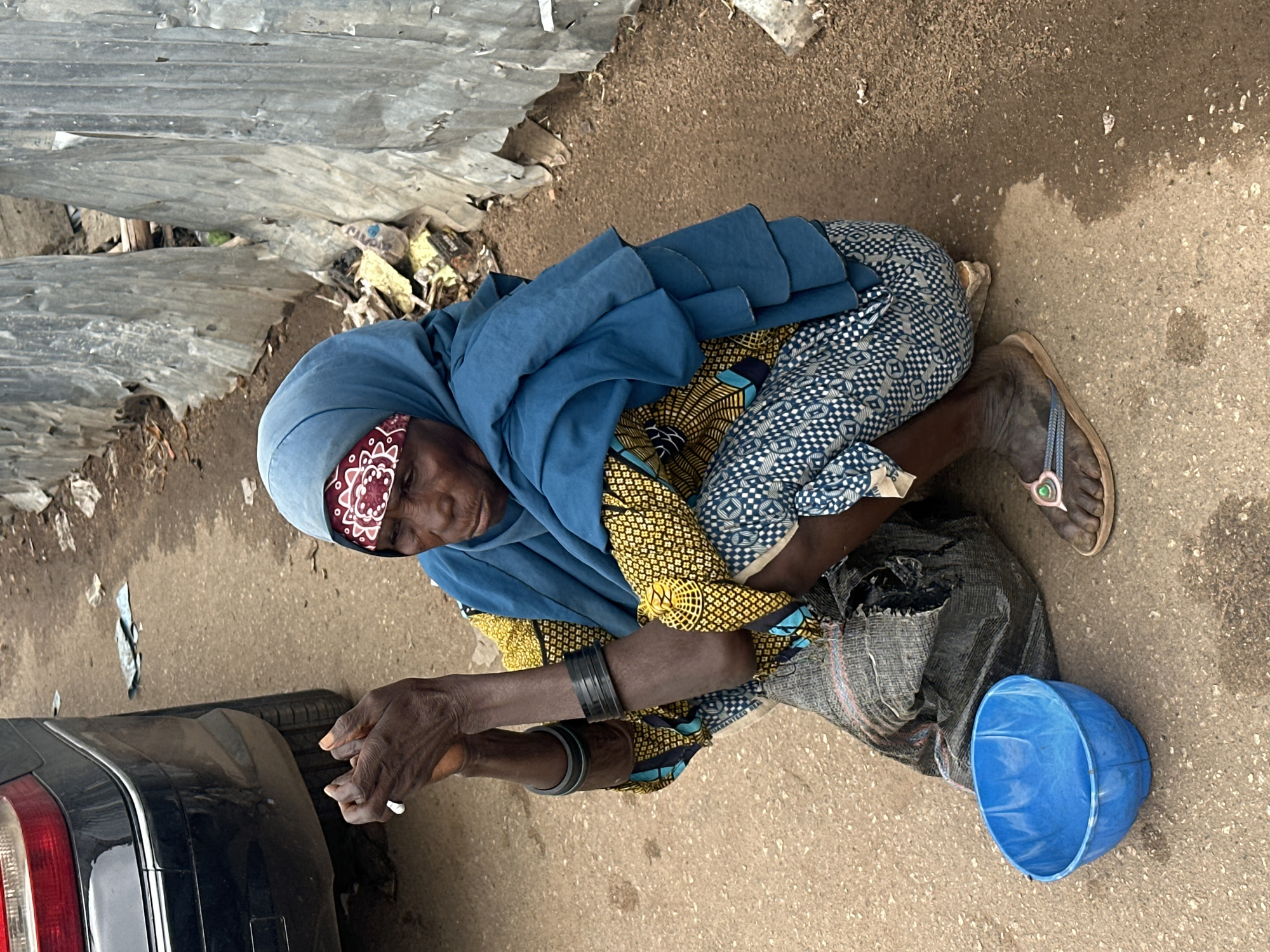 A Black woman with dark skin, wearing a blue headscarf and a patterned yellow and blue garment, kneels on a dirt and asphalt surface in Jos, Nigeria. She holds a cigarette in her hands, and wears dark bracelets on her arms. A blue plastic bowl rests on the ground in front of her, and a dark, torn bag is behind her. The background features a black car, a corrugated metal fence, and debris. The scene appears to be an outdoor, possibly urban environment, given the presence of the car and the man-made structures.