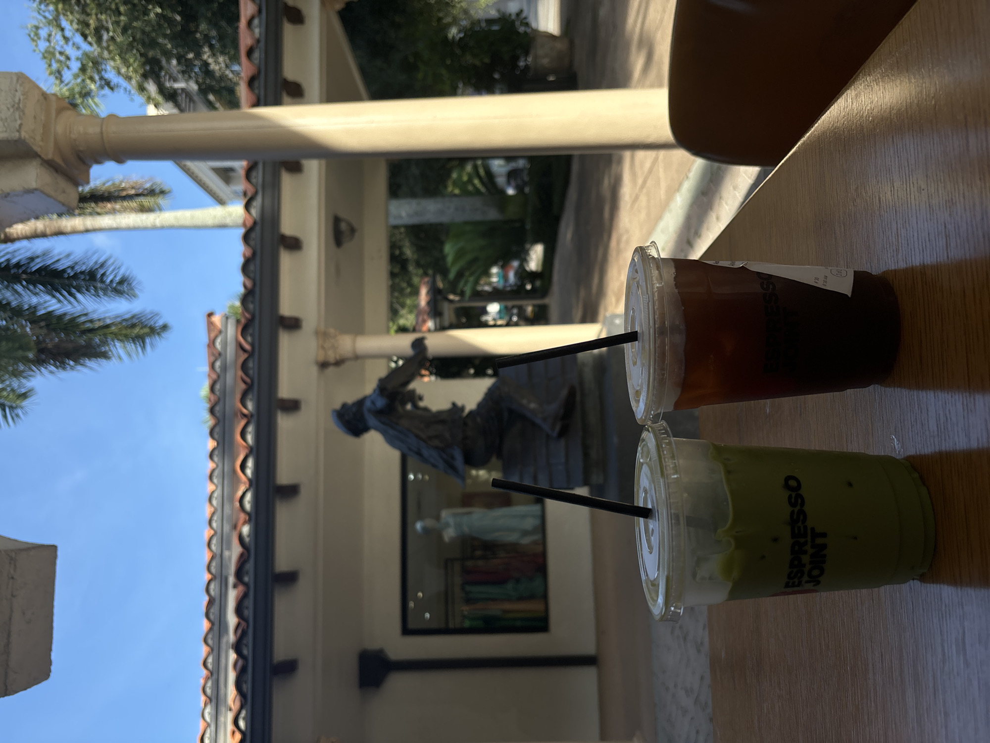 Two iced drinks are on a wooden table. The drink on the left is green and has "ESPRESSO JOINT" written on the cup. The drink on the right is dark brown and has a white sticker on the cup. Both drinks have black straws. In the background, there is a statue of a man sitting on a roof. Behind the statue is a store window displaying colorful clothing on mannequins. There are palm trees and blue sky visible. The scene appears to be outdoors.