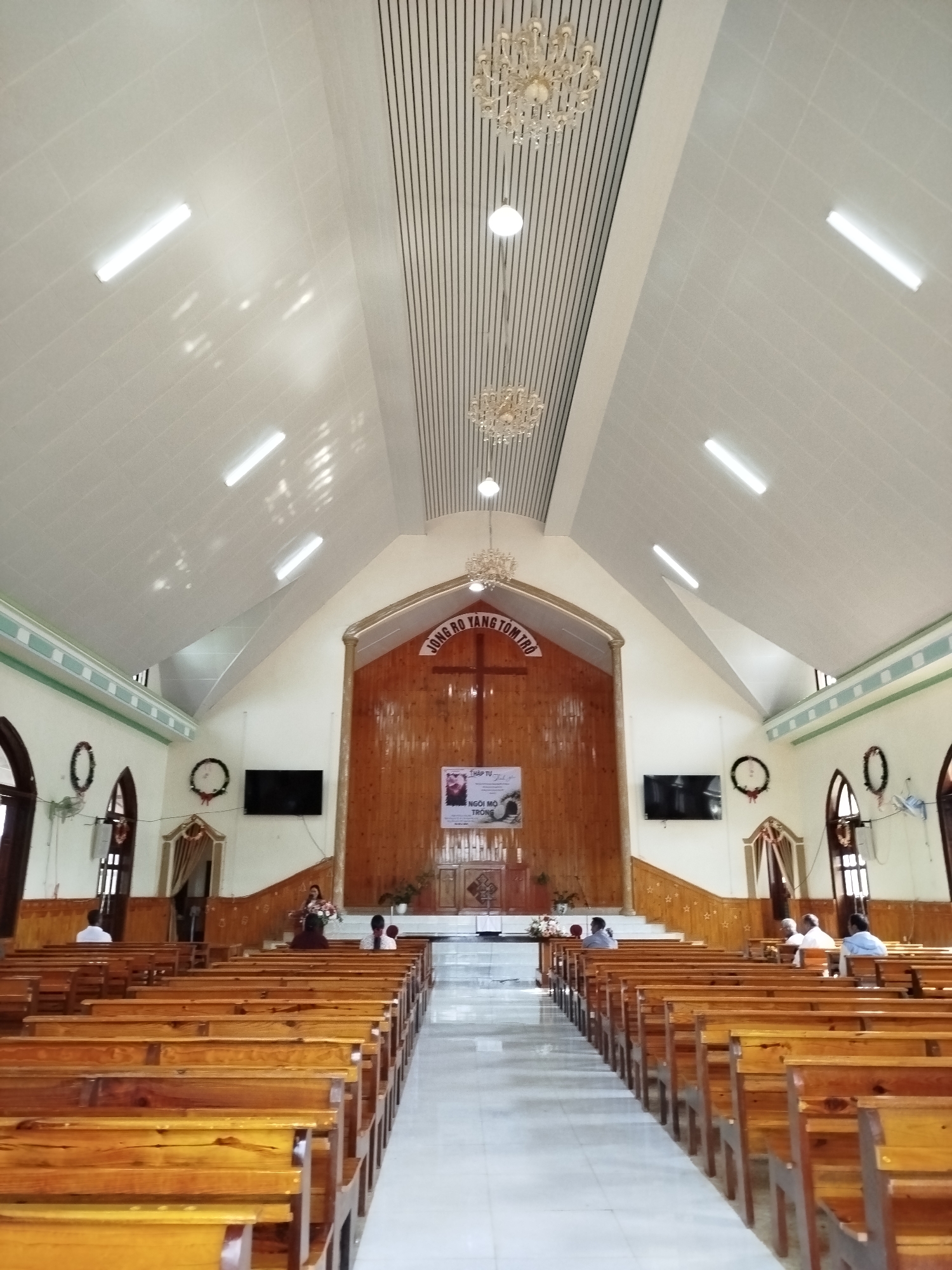 The image depicts the interior of a church in B'Liang, Vietnam. Rows of wooden pews are visible on either side of a central aisle leading to the altar. Several people are seated in the pews. The altar is a wooden structure adorned with a cross and a banner. Two television screens flank the altar. The church has a high ceiling with linear lighting and chandeliers. The walls are white with green trim. Windows and decorative wreaths are visible along the walls.