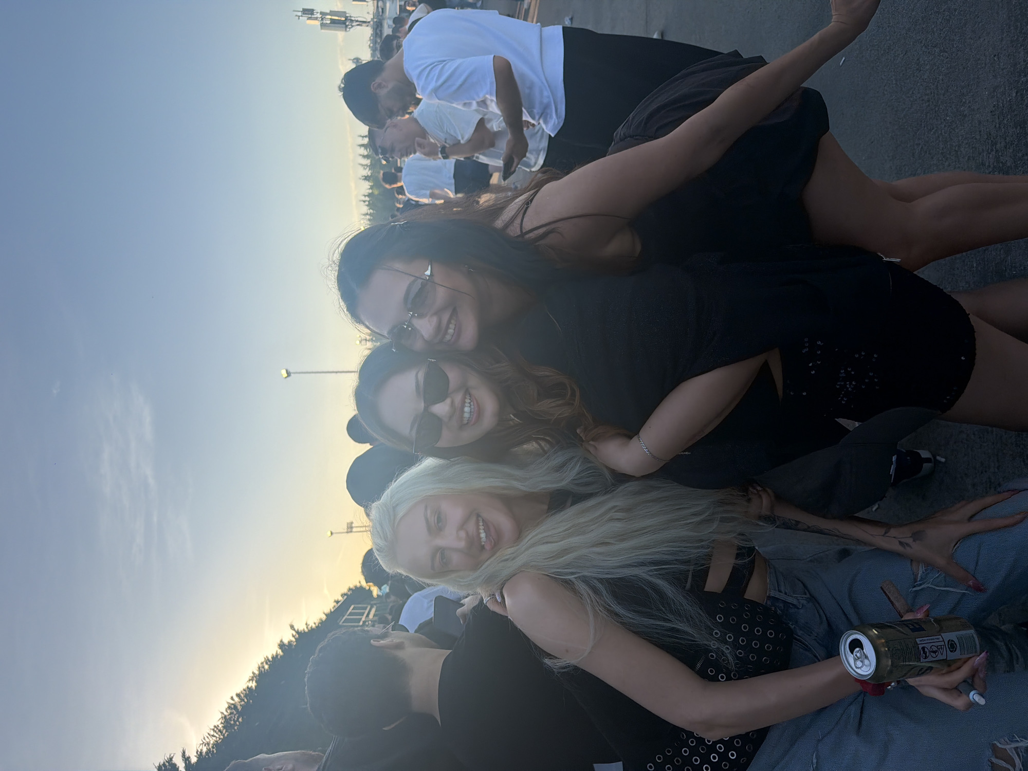 A photograph shows three women posing together outdoors in what appears to be a public space with a crowd of people. The woman at the bottom of the frame has long blonde hair, is wearing a black top with silver eyelets, and is holding an open can of what appears to be an alcoholic beverage. Her left hand is tattooed. She is seated and wearing denim pants. Above her is a woman with sunglasses and reddish-brown hair. At the top of the frame is a woman with long dark hair and sunglasses, wearing a black dress. All three women are smiling. The background shows additional people, some buildings, and a sky with a sunset effect. The location is potentially Başakşehir, Turkey.