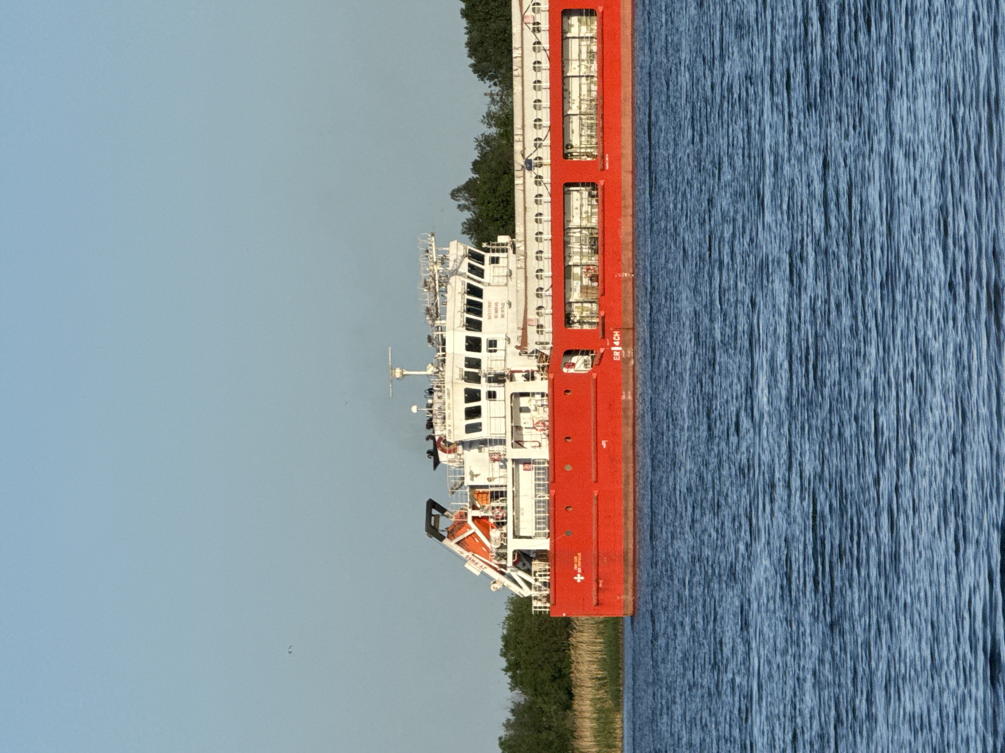 A red and white ship is situated in a body of water. A grassy bank with trees borders the shoreline. The sky is clear. The location is likely Rostov-on-Don, Russia, based on context.