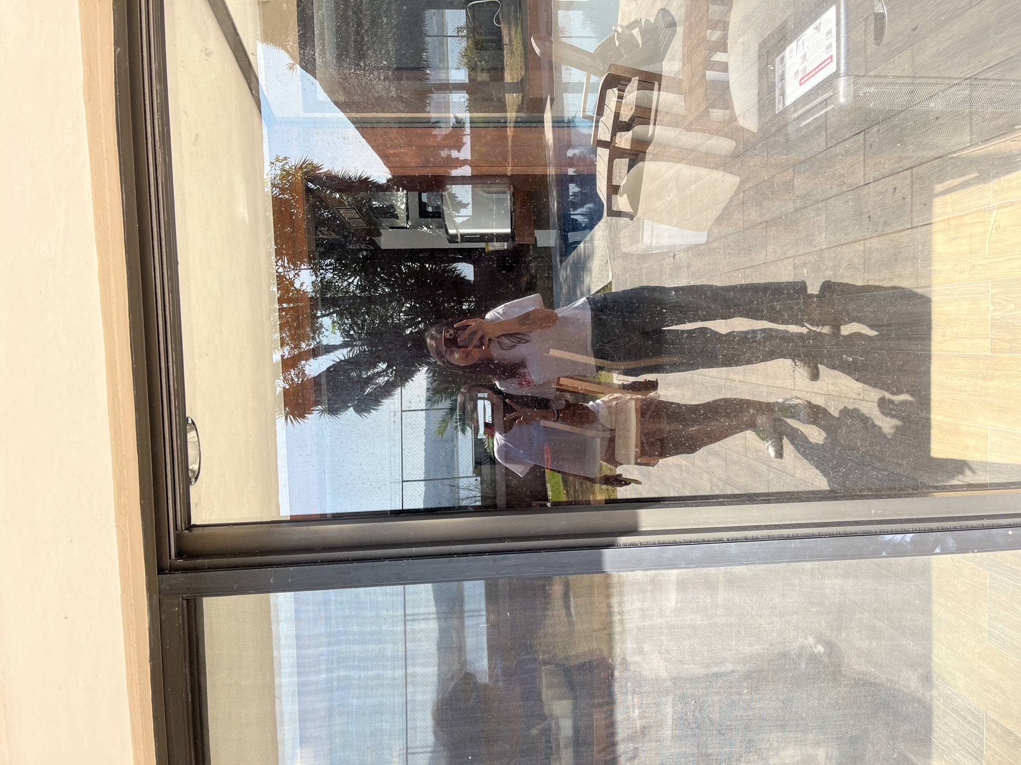 The image shows a reflection in a window in El Oasis Valsequillo, Mexico. The reflection includes a person holding a mobile phone, palm trees, a building, and outdoor furniture. The foreground shows the window frame and partially obscured view of the outdoors through the window. The person appears to be taking a picture of themselves in the reflection.