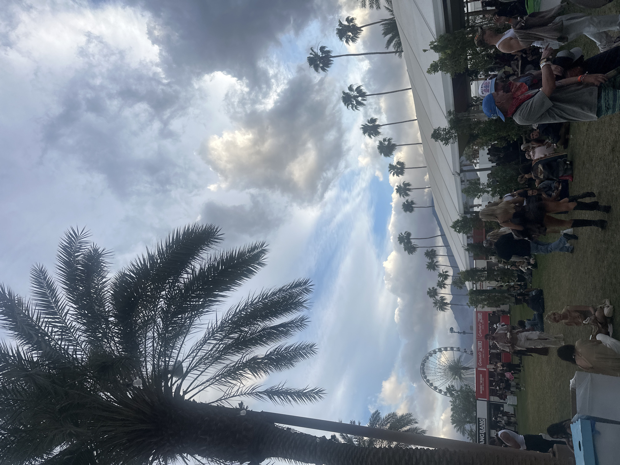 The image captures an outdoor event in what appears to be a city, possibly in a desert environment given the presence of palm trees. A large Ferris wheel is visible in the background, suggesting a carnival or fair atmosphere. There are crowds of people, some of whom are casually dressed, suggesting a relaxed, daytime event. There are tents or structures nearby. The sky is partly cloudy.