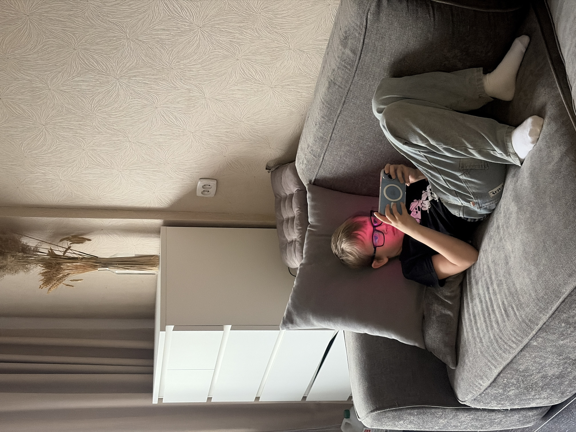 A young person lies on a grey fabric couch, wearing a black shirt with a cartoon design and grey denim pants. They are holding a blue cell phone with a round camera lens housing close to their face. The person is wearing glasses and has blonde hair. The couch is positioned against a wall with a decorative off-white wallpaper. To the left, a white dresser with slanted drawer fronts is visible. A dry floral arrangement sits on top of the dresser. The setting appears to be indoors. The location in Saint Petersburg, Russia, is not discernible from the image.