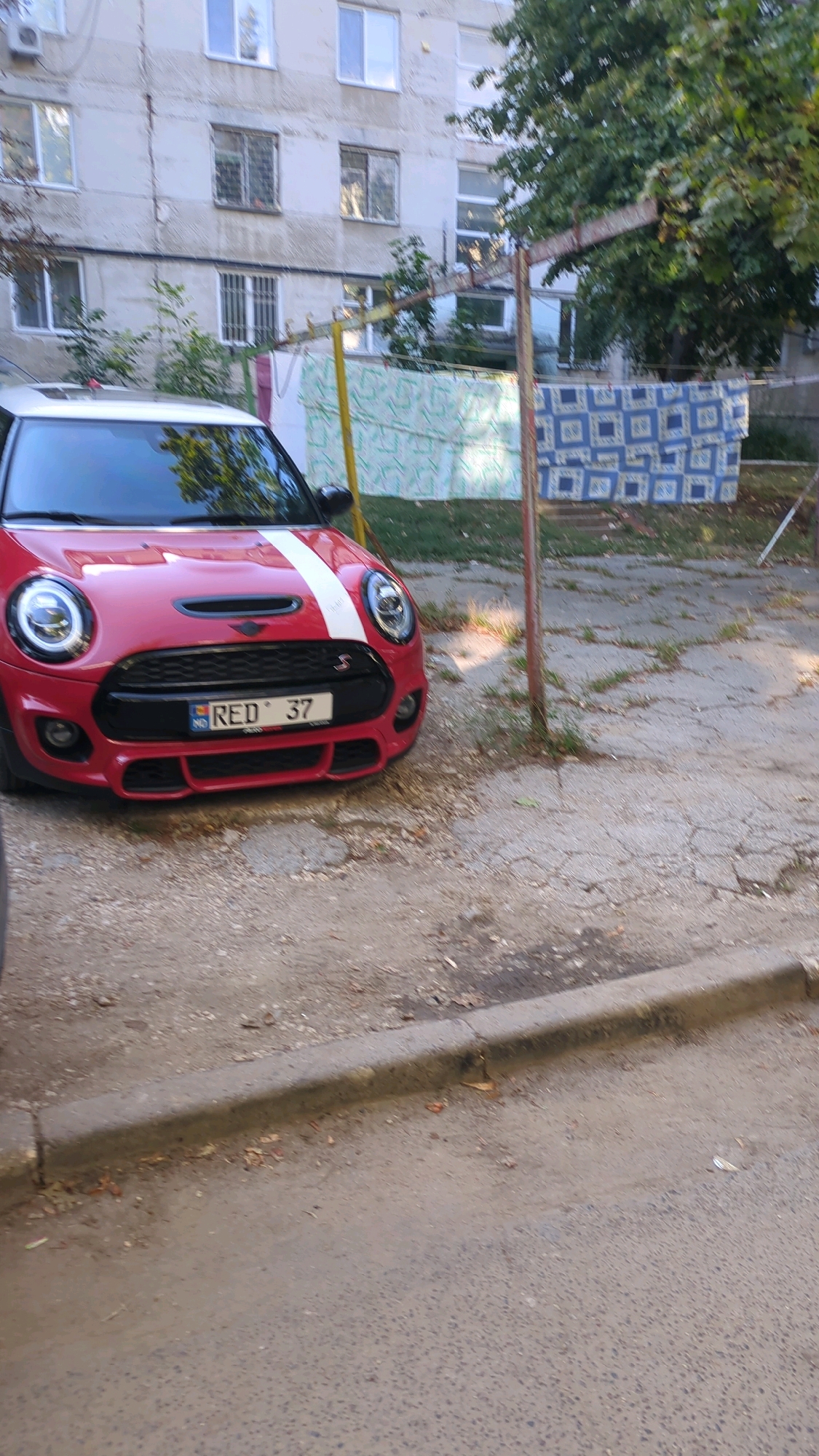 A red Mini Cooper with a white stripe down the hood and roof is parked on a cracked asphalt surface in front of a light-colored, multi-story building. The car's license plate is visible and reads "RED 37". In the background, clothes are hanging on a clothesline strung between a metal frame and a pole. The building appears to be an apartment complex with multiple windows. Trees are visible behind the clothesline. The ground around the car is covered with gravel and fallen leaves.