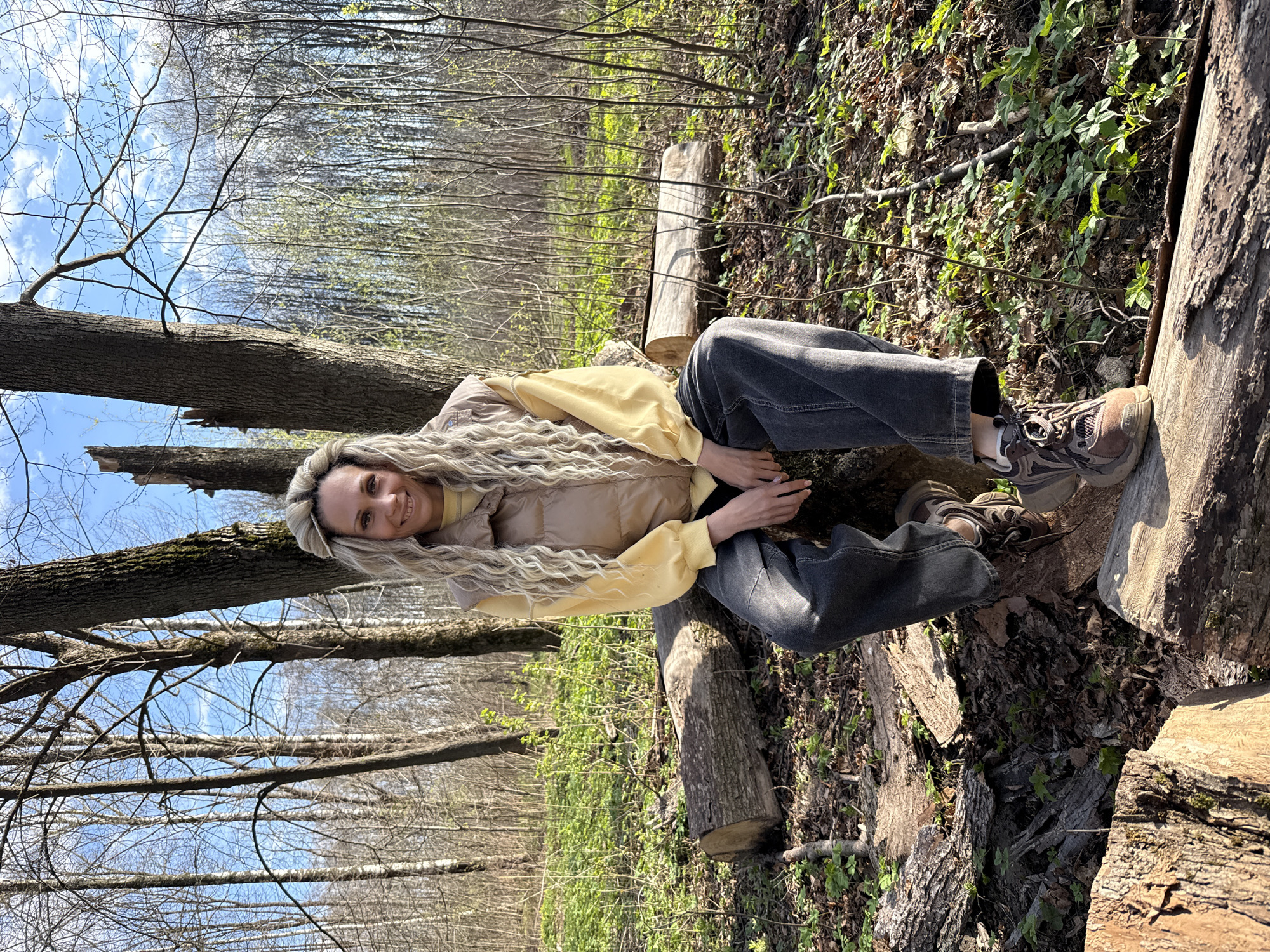 A young woman is sitting on a fallen log in a wooded area. She is wearing a light yellow long-sleeved shirt, a beige vest, grey jeans, and brown sneakers. She has long blonde hair and is smiling at the camera. The background shows trees and greenery, with a blue sky visible through the branches. The location is the forest. Rusanovo, Russia, is not visually identifiable from this image.