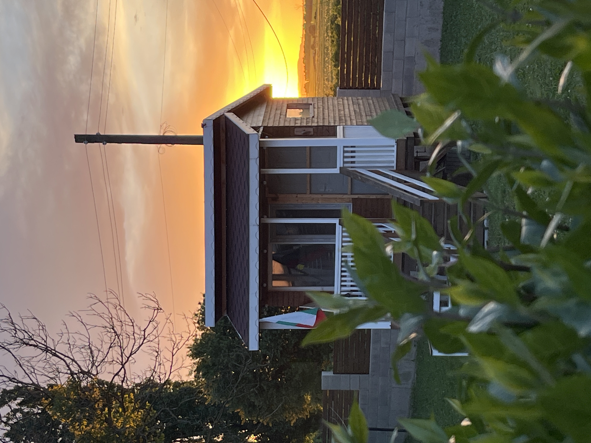 Captured at dusk in Pchelnik, Bulgaria, the image presents a one-story building with a dark-shingled roof and white-railed porch. A Bulgarian flag is visible attached to the building. The setting sun creates a bright, orange sky visible behind the structure. High-voltage power lines stretch across the sky. In the foreground, green foliage partially obscures the view. A wooden fence borders the property. A tree with bare branches is visible on the left.