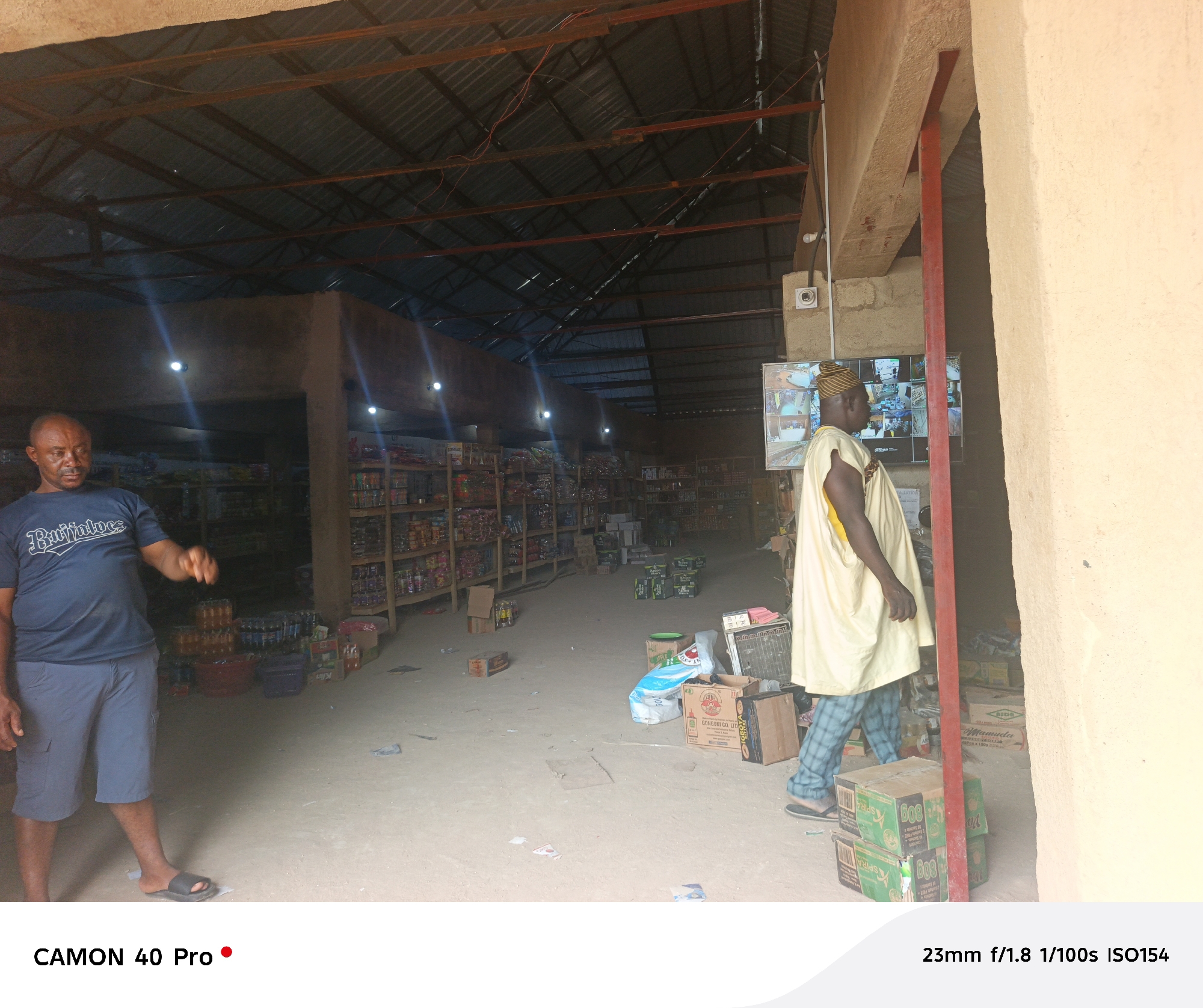 The image shows the interior of a general store in Langtang, Nigeria. The store has a corrugated metal roof supported by beams, with visible wiring. Fluorescent lights are attached to the walls, illuminating the shelves stocked with various items. A dark-skinned man in a blue t-shirt and shorts stands on the left. On the right, another man wearing a light-colored traditional outfit with a striped hat walks past a doorway. Behind him, there is a security monitor displaying multiple camera feeds. Boxes and various items are scattered on the dirt floor. There are shelves stocked with goods in the background. The boxes nearest the camera on the right include labels like "GONGONI CO. LTD" and "Mamuda".