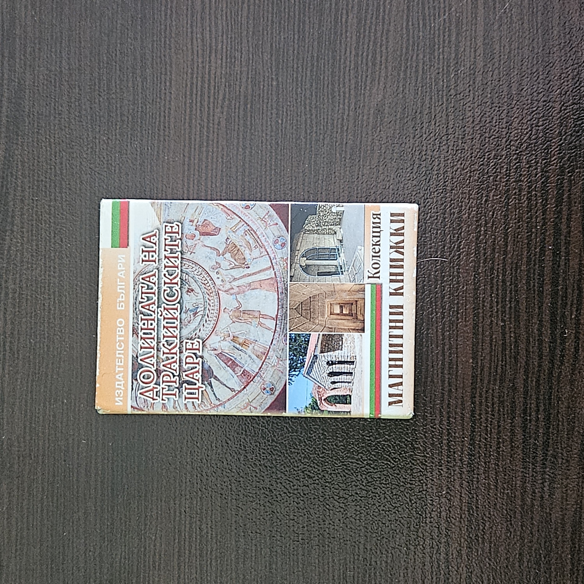Here is a description of the image:
The image shows a small, rectangular book resting on a dark, textured surface. The book has a cover featuring Bulgarian text and images related to Thracian history. A tricolor flag (white, green, red) runs vertically on both the left and right sides of the cover. The book's title, partially visible, appears to be "ДОЛИНАТА НА ТРАКИЙСКИТЕ ЦАРЕ", likely referring to the Valley of the Thracian Kings near Stara Zagora, Bulgaria. The cover's design incorporates a circular image resembling an ancient fresco or mosaic, along with photographs of architectural structures, possibly from Thracian tombs or sites in the area. Other text in Bulgarian reads "ИЗДАТЕЛСТВО БЪЛГАРИ" and "Колекция МАГНИТНИ КНИЖКИ". This likely refers to a Bulgarian publishing house producing a series of miniature books.
This image strongly suggests the book is a tourist or educational publication about Thracian history and culture, particularly focusing on the area surrounding the Valley of the Thracian Kings near Stara Zagora.