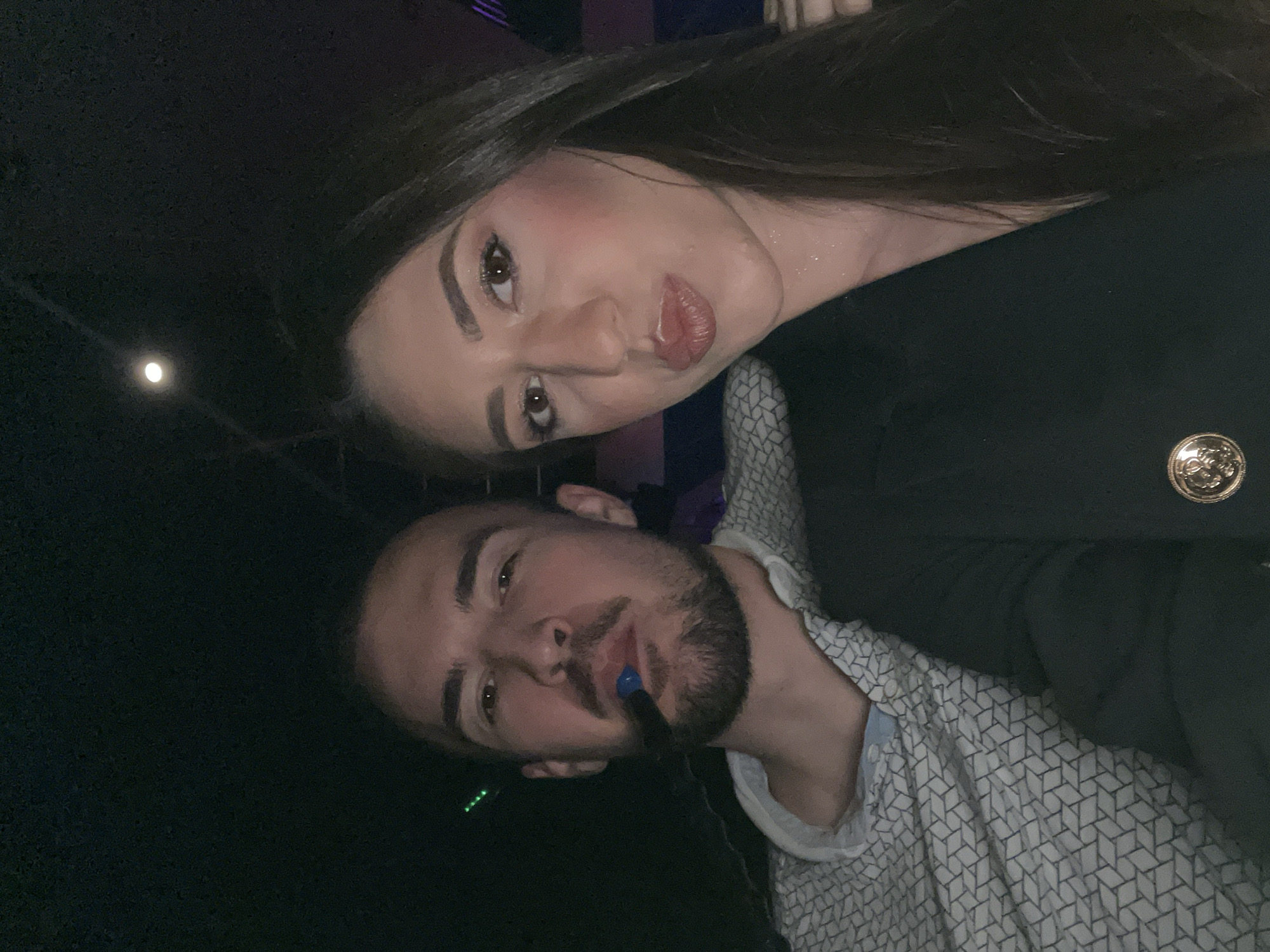 A woman and a man are posing for a selfie in a dimly lit location. The woman has long dark hair, fair skin, and is wearing a black blazer with a gold button visible on the right side. She is making a puckered lip face. The man has a trimmed beard and is wearing a patterned white and grey shirt. He is holding what appears to be a small object in his mouth. The background is dark, with a few indistinct light sources. There is no indication of Sofia, Bulgaria.
