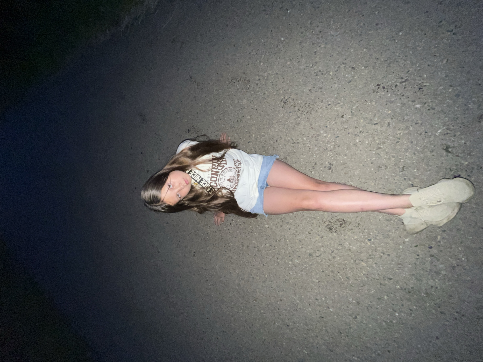 A young girl with long, light brown hair is sitting on an asphalt road at night. She is wearing a white t-shirt with graphics and light-colored denim shorts. Her legs are crossed, and she is wearing light-colored sneakers. Her head is tilted, and she is looking towards the camera. The road appears to be a rural road, possibly a country lane. The background is dark, suggesting it is nighttime. There is no specific indication of the city of Novopokrovskaya, Russia.