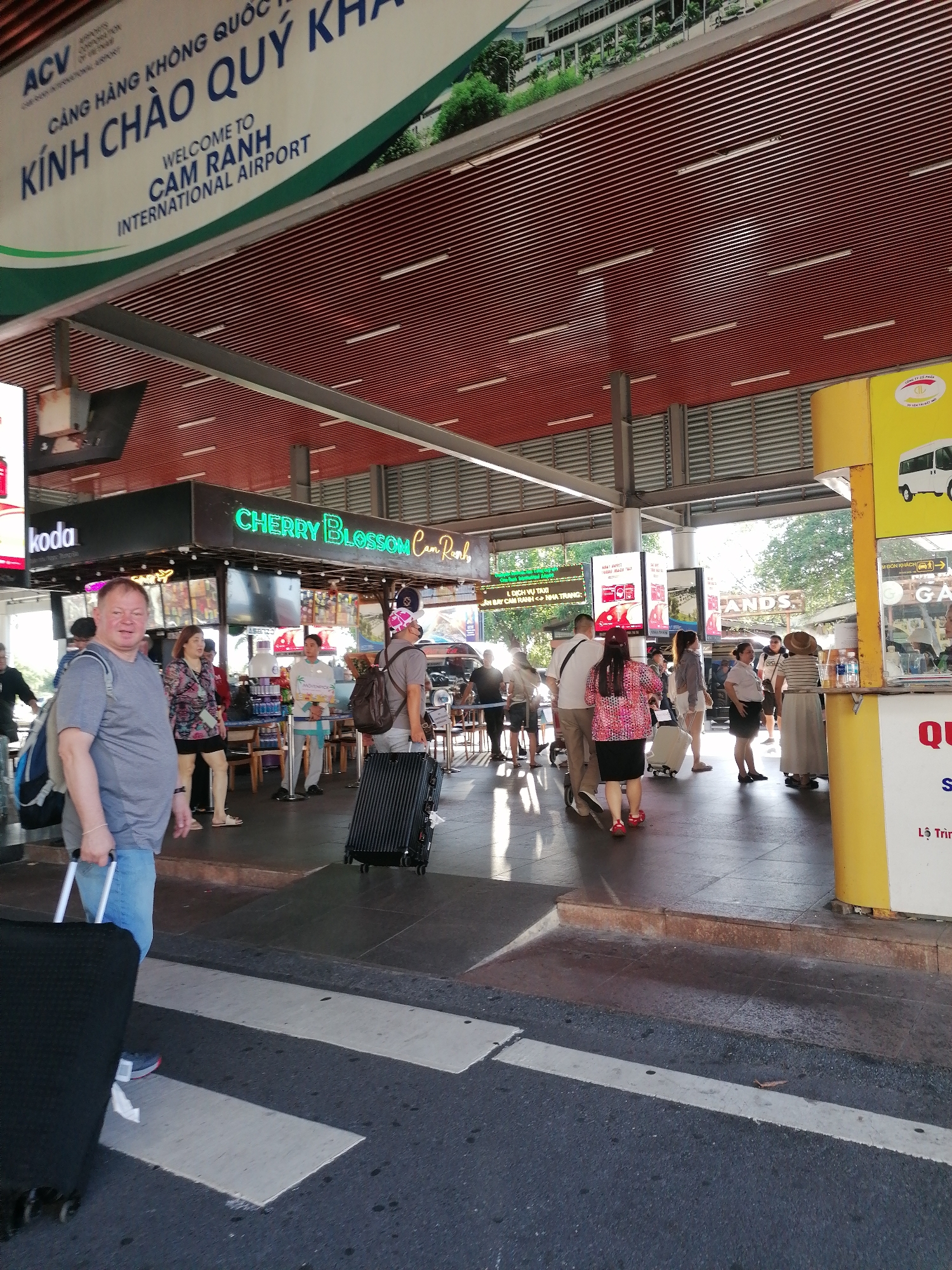 The image captures a scene at Cam Ranh International Airport in Cam Ranh Ward, Vietnam. Overhead, a sign reads "Welcome to Cam Ranh International Airport". People are standing on a covered walkway outside the airport. Some are waiting near a yellow taxi stand, while others are gathered near a Cherry Blossom cafe. The ground is marked with a zebra crossing. A man in the foreground pulls a suitcase.