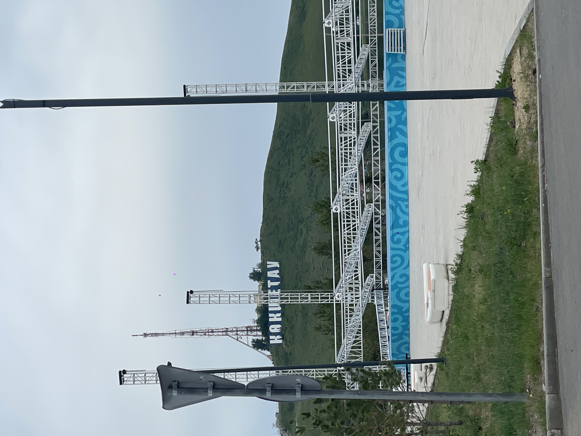 The image shows a view in Kokshetau, Kazakhstan. A green hillside is visible in the background, with the name of the city, "КОКШЕТАУ", displayed in large, white, vertically oriented letters. A series of white metal structures stand in the foreground, possibly related to event staging or decoration. These structures have a framework appearance. There is a blue patterned design on the ground. A black lamppost and road signs are in the lower part of the image. The sky is overcast. A concrete path runs next to an area of grass. A road is at the extreme right edge of the frame. There are communication towers visible in the background.
