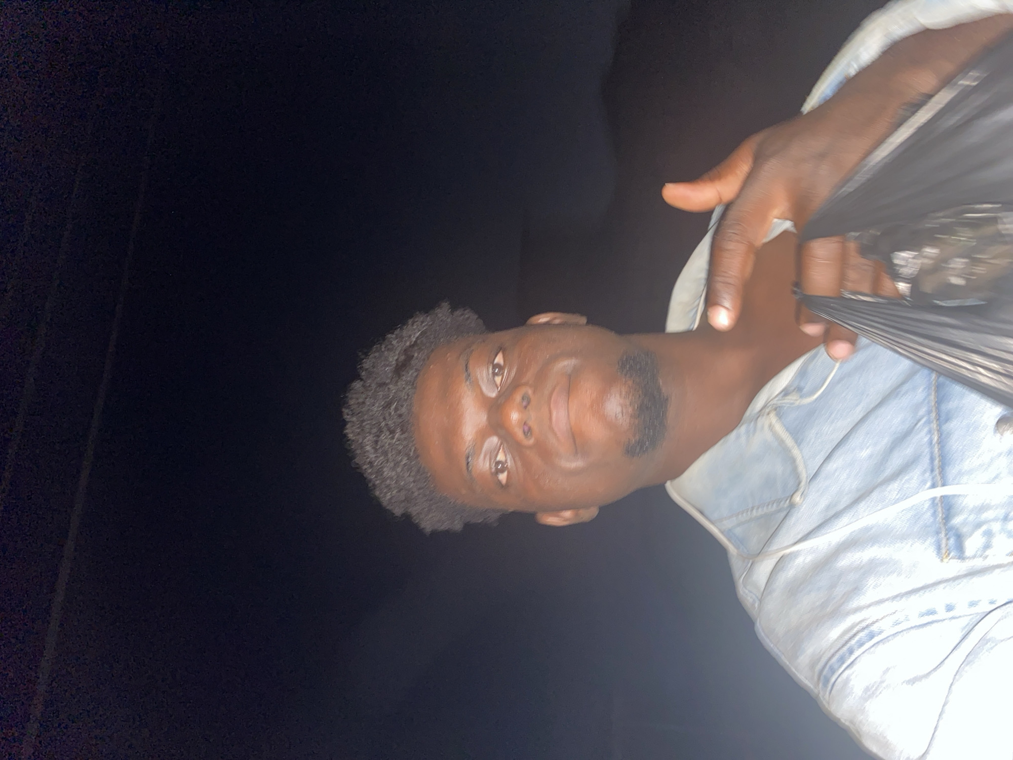 This media file is a low-light portrait of a young man, likely a selfie, taken in a dark environment, suggesting nighttime. The image is rotated counter-clockwise.
The primary subject is a man with dark skin, short, curly black hair, and a neatly trimmed goatee. He is looking directly at the camera with a neutral to slightly smiling expression. He is wearing a light-colored, possibly light blue or faded denim, jacket or shirt. A thin, white cord or string is visible around his neck area, potentially part of his clothing or a necklace. His right hand is raised and visible, holding a dark, pleated object that appears to be a black plastic bag or a folded fan.
The background is almost entirely black, with minimal discernable detail, reinforcing the impression of a very dark setting, such as outdoors at night or a dimly lit indoor space. The subject is illuminated by a direct light source, possibly a camera flash, which creates a high contrast between him and the dark background. No specific location cues or visible text are present in the image.