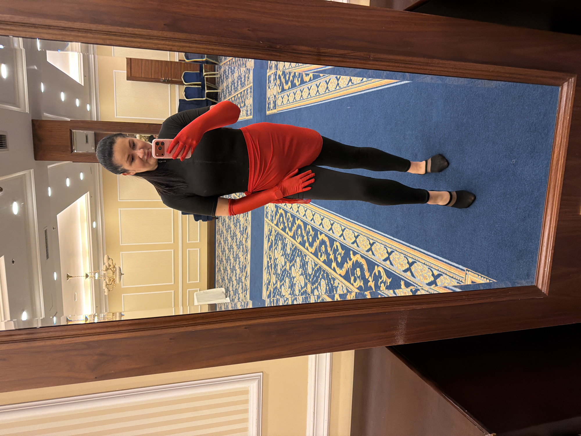 A woman is taking a mirror selfie in a room with a blue carpet and yellow walls. She is wearing a black top, black leggings, and a red wrap skirt. She also has on red gloves and black sheer pointed-toe heels. The room has a chandelier and chairs in the background.