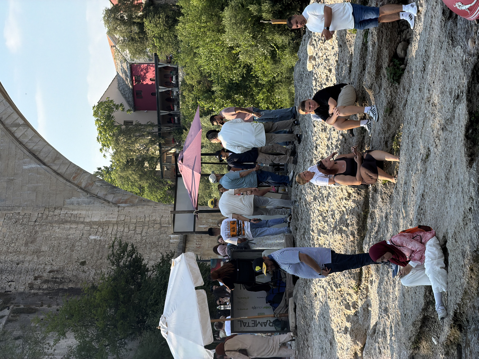 This is a vertical, eye-level shot of a group of people gathered on a rocky embankment overlooking a river, with a stone bridge and buildings in the background. The setting appears to be Mostar, Bosnia and Herzegovina, given the presence of a structure resembling the Old Bridge.
The people are engaged in various activities. Some are standing and talking, others are seated, and one person is lying down. There's a woman in the foreground wearing a black tank top and skirt, lying on the rocks and looking towards the camera. Next to her, a man in a white t-shirt and blue shorts is seated and holding a phone. Further up the embankment, a group of men are standing and conversing. One man in a white t-shirt is looking at his phone, while another in a black t-shirt and beige shorts is seated. A woman in a pink and white patterned top and white pants, wearing a burgundy headscarf, is also seated and looking at her phone. There are several other individuals scattered throughout the scene.
Two large white umbrellas are set up on the embankment, suggesting a casual gathering or a place where refreshments might be served. One umbrella is fully open, while the other is partially visible. In the mid-ground, there is a pink umbrella over what appears to be an outdoor seating area with tables and chairs. Behind the people, a red building with a stone roof and arched windows is visible, along with lush green trees and a stone wall that seems to be part of an old fortification. A large stone archway, possibly part of the bridge, is visible on the left side of the image.
The lighting suggests it is daytime, likely late afternoon, with the sun casting warm light on the scene and creating shadows. The overall mood seems relaxed and social, with people enjoying the view and each other's company.
Visible text includes "RAMETAL" on a sign near the umbrellas, and some smaller text on a sign further back, which is difficult to read clearly.