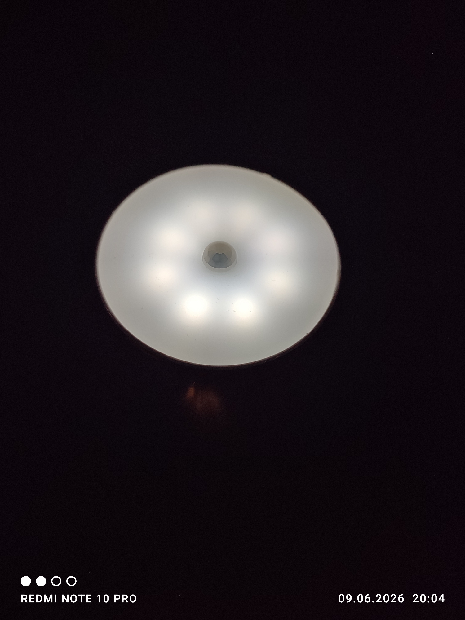 An indoor ceiling light fixture is depicted in the center of the frame against a dark background. The fixture is circular with a frosted diffuser covering multiple LED bulbs, which are illuminated. A small, multifaceted sensor is visible in the center of the fixture. The bottom left of the image contains the text "REDMI NOTE 10 PRO" and four white circles. The bottom right corner displays the date "09.06.2026" and the time "20:04". The overall scene is dark, with the light fixture being the primary source of illumination.