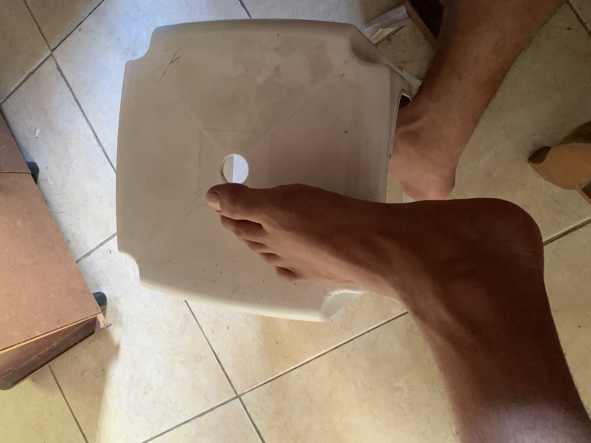 The image shows a person's feet and lower legs resting on a white plastic stool in what appears to be an indoor environment with tiled flooring. The stool has a circular opening in its center. In the background, a wooden cabinet with wheels is visible. The overall scene suggests a casual, everyday setting, possibly a home or a utility area. The lighting indicates it is daytime. The provided location, Santa Ana, Venezuela, is contextual information that cannot be verified solely from the visual content of the image.