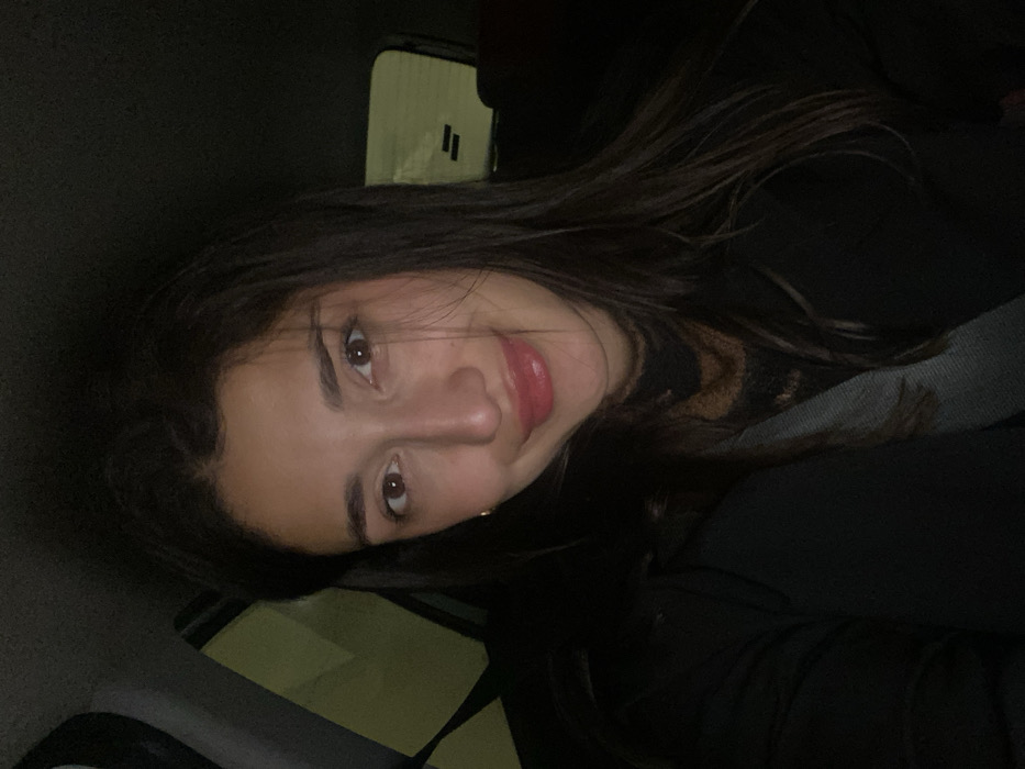 The scene depicts a female individual inside a vehicle, illuminated by an immediate light source, likely a camera flash or phone screen, against a dark, low-light background. The precise location within the city of Đurmani, Montenegro, cannot be determined from the visual information provided.
The primary subject is a young adult female, positioned centrally, facing directly towards the camera. She possesses long, dark brown hair, parted on the left, with some strands falling over her forehead and shoulders. Her complexion is olive, and she has brown eyes. A faint gold hoop earring is visible on her left earlobe. Her lips are coated with a glossy, light pink substance, and she displays a slight, closed-mouth smile. She is wearing a dark-colored, possibly black, outer garment over a dark, textured or patterned high-neck top. A grey seatbelt diagonally traverses her torso from her left shoulder towards her right hip.
The immediate environment appears to be the interior of an automobile. Blurred light sources, predominantly yellow-green, are visible through the vehicle's windows on the left side, suggesting an outdoor setting at night or in a poorly lit area. The rear window is visible behind her right shoulder, showing indistinct dark shapes and faint horizontal lines, potentially elements of a defroster or reflections. The vehicle's interior surfaces are dark and indistinct due to low light. No other individuals or specific objects are clearly discernible in the background.