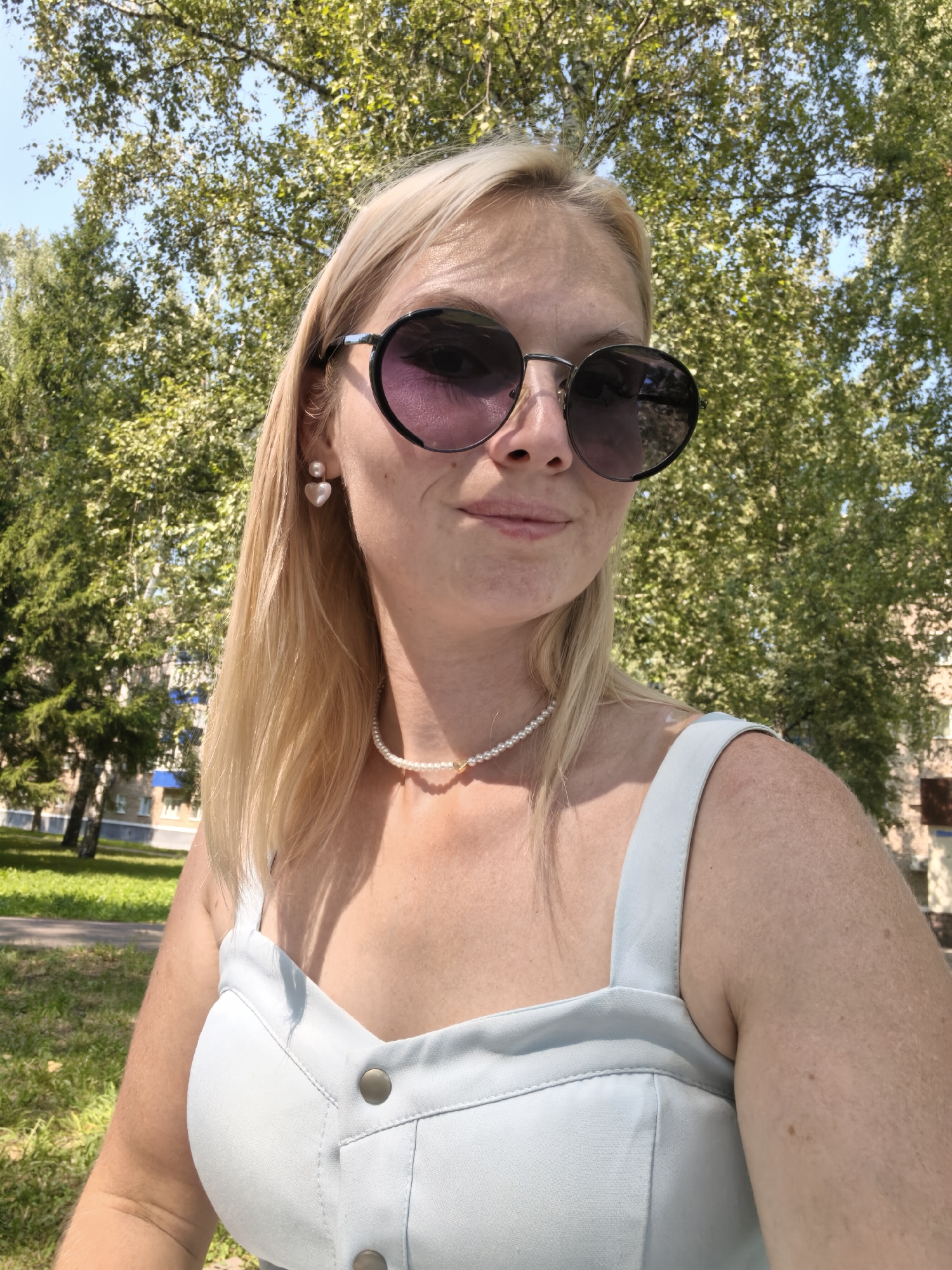 In Sterlitamak, Russia, a blonde woman with fair skin and freckles takes a selfie outdoors on a sunny day. She is wearing large, round, dark sunglasses with purple lenses, a pearl necklace, and a light blue, button-front tank top. Her hair is shoulder-length and blonde. She has pearl earrings shaped like hearts. The background consists of lush green trees, foliage, a patch of grass, and a building with a blue roof and white walls. The lighting suggests a midday or early afternoon time.