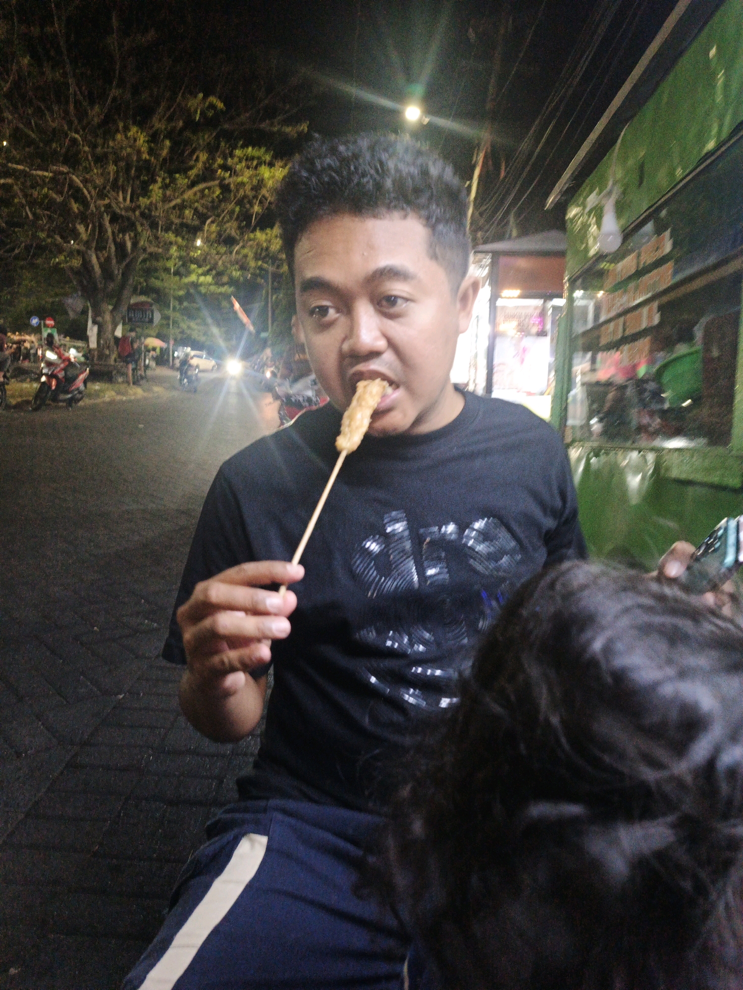 A medium shot captures a young man in Surabaya, Indonesia, eating a piece of food on a skewer. He is wearing a black t-shirt and blue shorts with a white stripe down the side. The man is in focus, with a blurry background that shows a street scene at night. There are trees, streetlights, and passing vehicles, including motorcycles. To the right of the man, a green food stall is visible. In the foreground, the back of another person's head is partially visible. The image appears to be taken in an outdoor, public area, likely a street or sidewalk in a bustling part of the city.