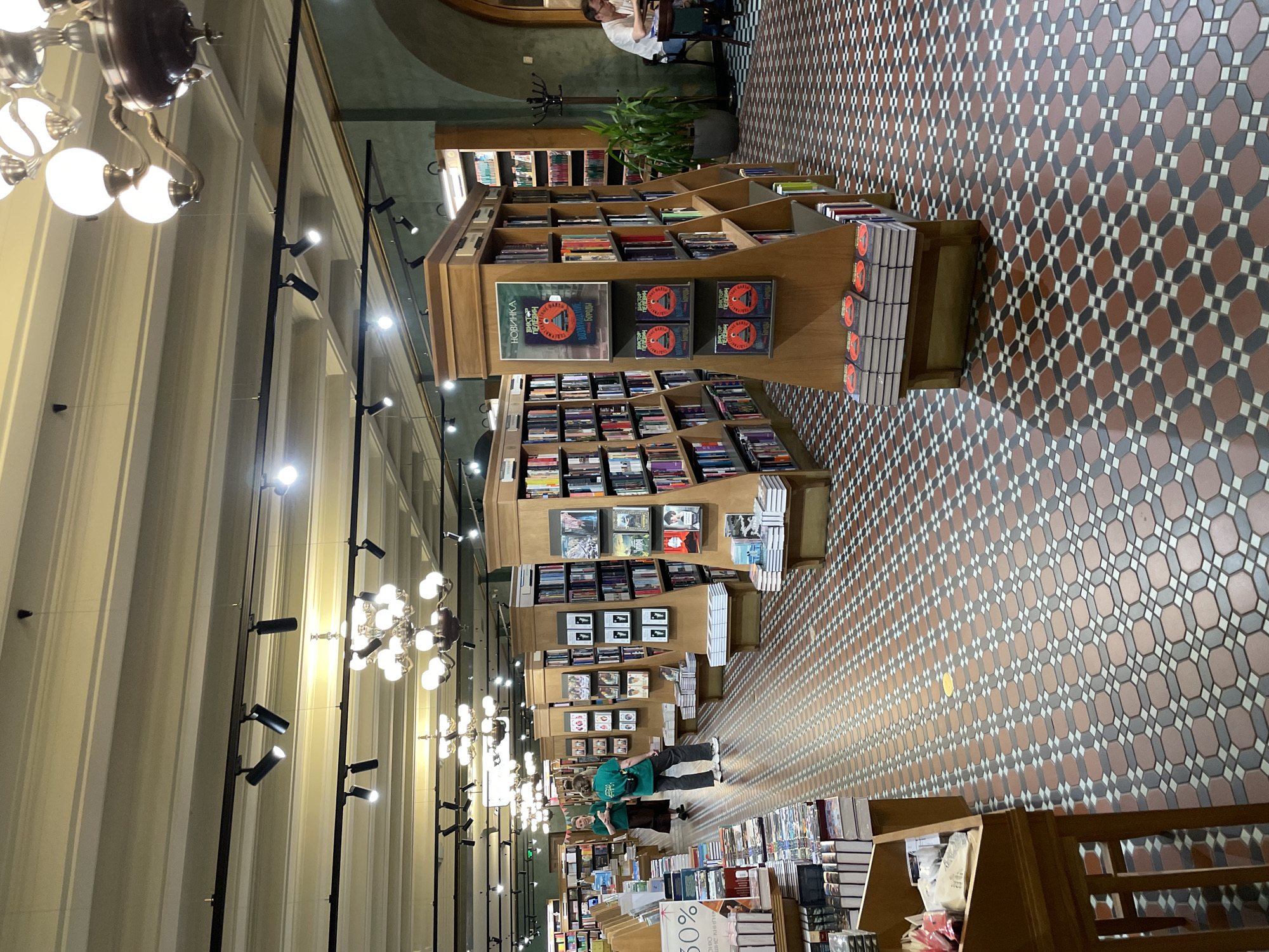 This detailed interior image depicts the Dom Knigi (Singer House) bookstore located on Nevsky Prospekt in Saint Petersburg, Russia.
The scene presents a wide view of the bookstore's main floor, characterized by a distinctive geometric tiled floor pattern consisting of octagonal reddish-brown, dark grey, and small white square tiles arranged in a repeating mosaic. Numerous tall, light-brown wooden bookshelves are arranged in aisles, extending deep into the perspective. Many shelves are filled with books, and additional stacks of books are displayed on lower platforms and tables.
In the mid-ground, a prominent display stand features copies of "Возвращение бороды" (The Return of the Beard) by Viktor Pelevin, clearly marked with "НОВИНКА" (New Arrival) at the top. To the left, a display table shows a "30%" discount sign in Russian.
Several individuals are present:
* Two women wearing matching green shirts, possibly staff members, are visible on the left side of the central aisle. One is walking away from the camera, and the other stands further back, facing away.
* In the far right background, a man with light hair is seated at a round table, possibly within a café section of the bookstore, engrossed in what appears to be a book or device. A dark coat rack stands nearby, and a large potted plant is situated between the bookshelves and the seating area.
* Other blurred figures are visible further down the aisles.
The high ceiling features elaborate moldings and multiple lighting sources: several ornate, multi-bulb chandeliers with white spherical globes, and numerous modern black track lights with individual spotlights illuminating the displays. The upper walls are painted a muted green, while the ceiling is a lighter, off-white tone. The overall lighting is bright and warm. A segment of the patterned floor on the far left shows a small yellow circular marker.