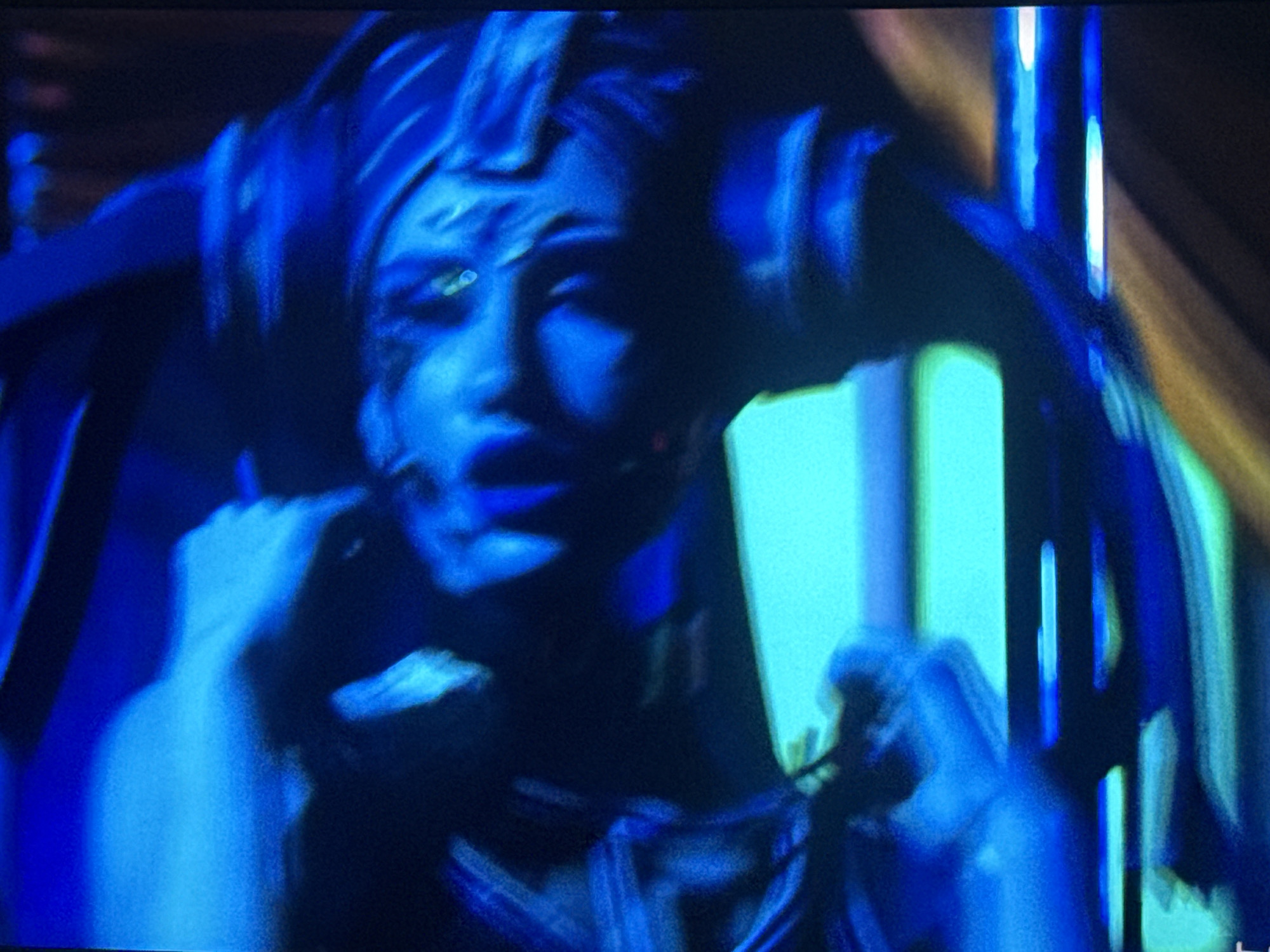 The image depicts a person with blue skin and what appears to be theatrical makeup, including markings around the eyes and possibly on the forehead. The person's lips are also blue. They are wearing a headpiece or elaborate hairstyle. The person is holding their hands up near their shoulders. The lighting is predominantly blue, with some yellow light in the background. The setting and location are undetermined.