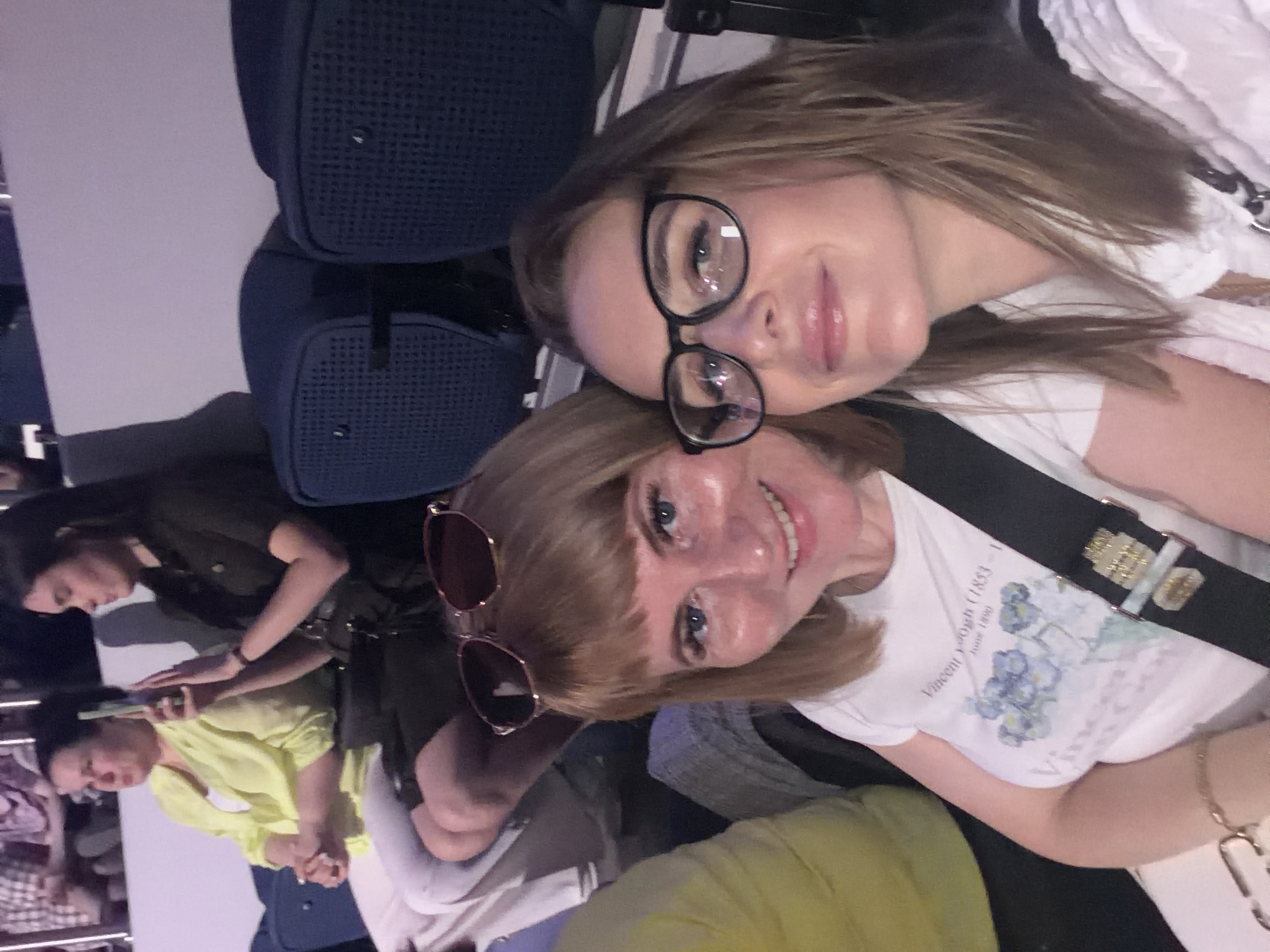 The image depicts two women taking a selfie in an indoor setting, likely an airplane. The woman in the foreground has short blonde hair with bangs and is wearing sunglasses perched on her head. She is smiling and wearing a white t-shirt with a floral design and a black strap across her chest. The woman in the background is wearing glasses and has shoulder-length brown hair. Behind them are two women seated, one wearing a yellow shirt and another in a green top using a cell phone. There are blue bags visible as well. The location appears to be inside an airplane.