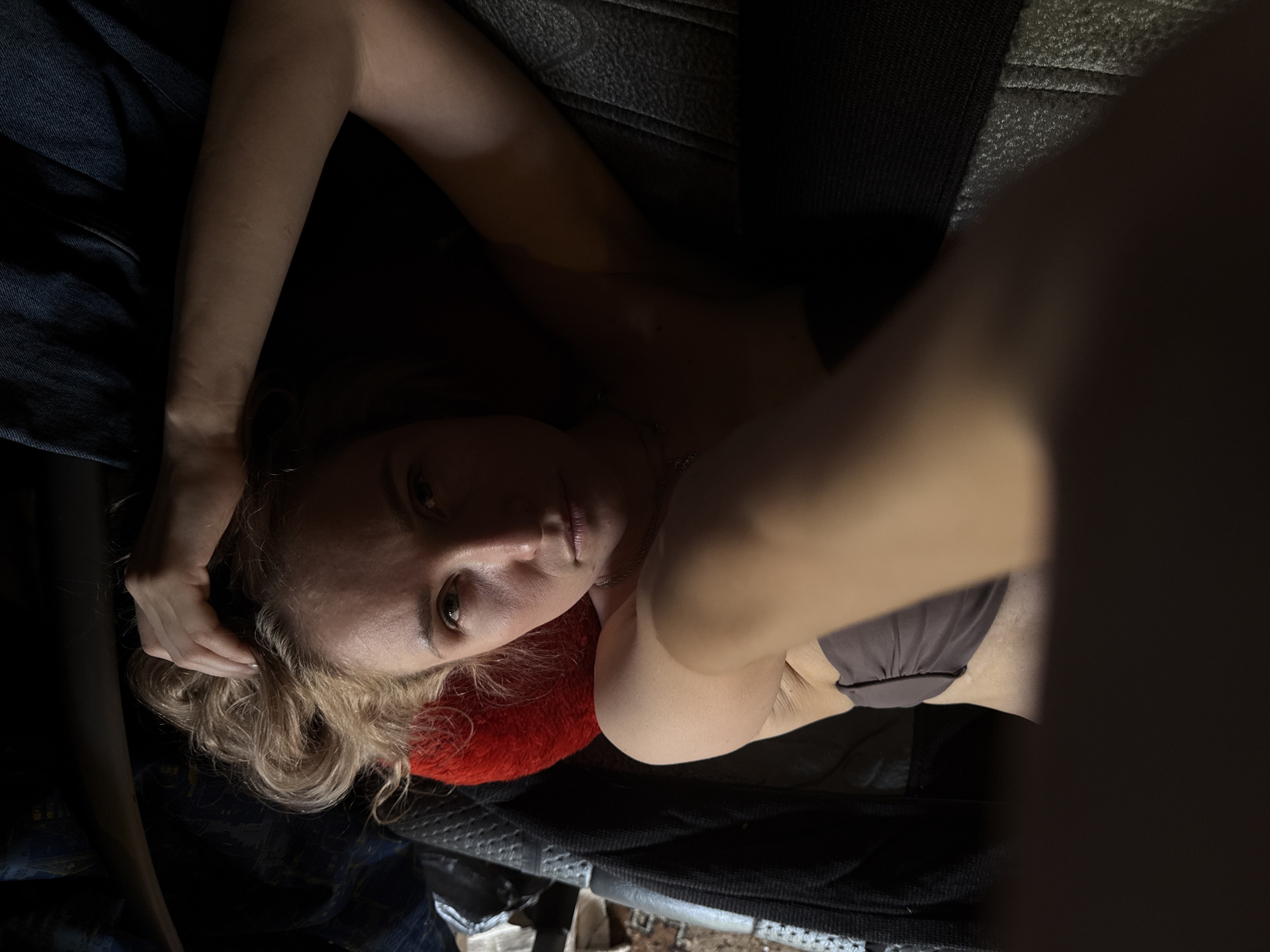 A woman is lying on a dark surface with a red pillow under her head. She is wearing a bra-like top. Her left arm is raised, with her hand near her head. The environment is dark and indistinct. The location is Усть-Тунтор, Russia.