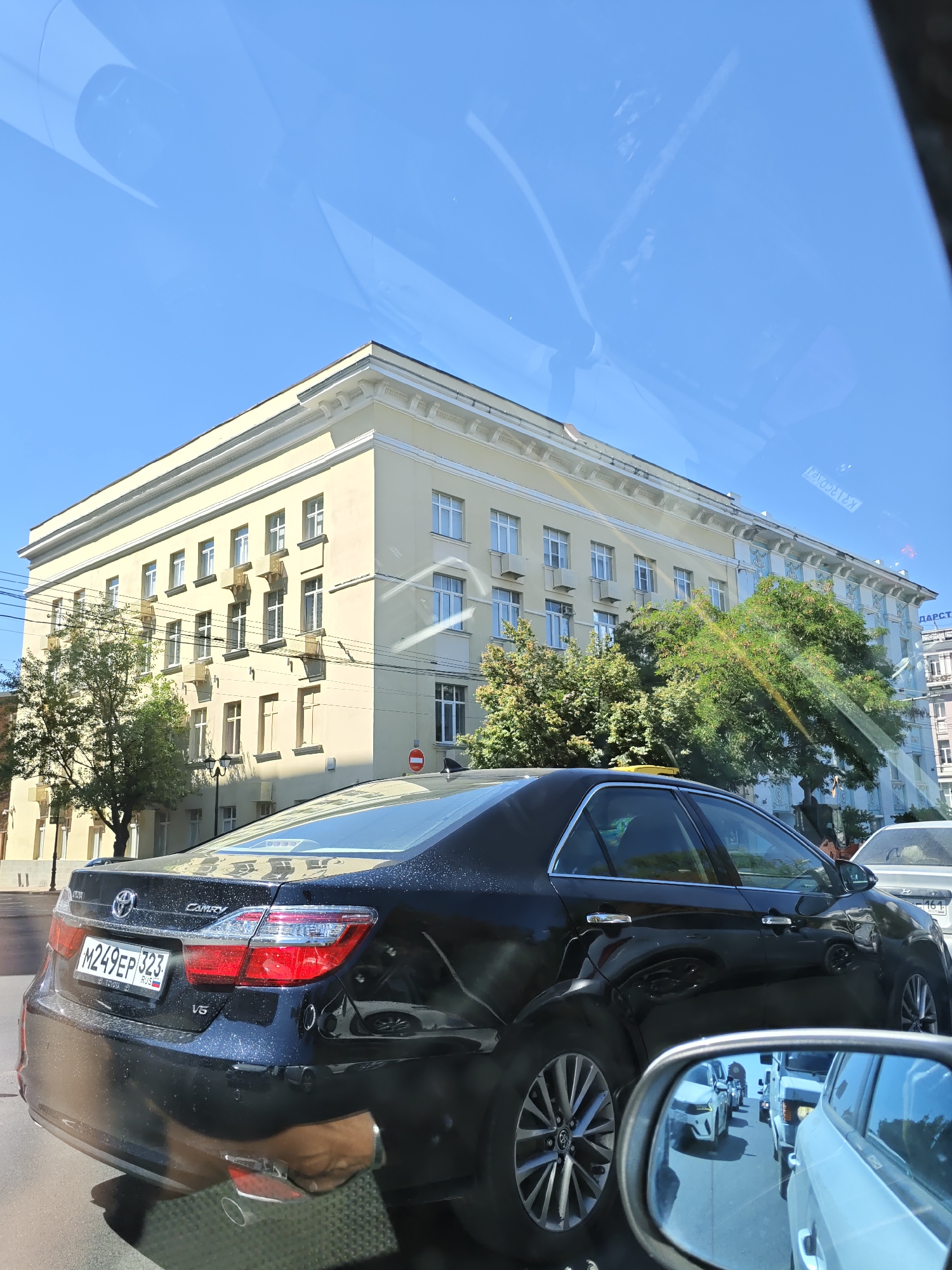 A black Toyota Camry, license plate M249EP 323 RUS, is parked on a street in Rostov-on-Don, Russia. The car is facing forward, and its rear end is visible in the foreground. In the background, a light yellow building with multiple windows and air conditioning units is visible. The building appears to be an office or residential building. Trees are also present, and the sky is clear and blue. The image appears to have been taken from inside another vehicle, as reflections of the sky and other vehicles are visible on the car's surface and in the side mirror. A sign with a red circle and a diagonal line is visible on the right side of the street, indicating a no-entry zone. The overall scene depicts a typical urban street in Russia on a sunny day.