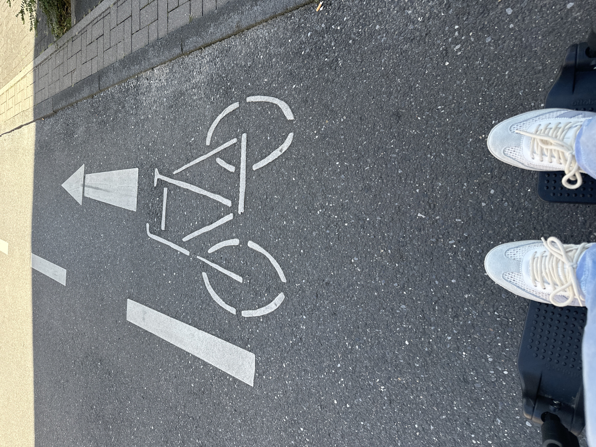 This is a first-person perspective, eye-level shot of a person on a mobility scooter, traveling on a marked bicycle lane in Krefeld, Germany. The asphalt surface of the lane is dark grey with visible aggregate. A white line, likely a lane marker, runs diagonally from the upper left towards the center of the frame. Further ahead on the lane is a white symbol depicting a bicycle, oriented to face forward, and a white arrow indicating the direction of travel. To the right of the bicycle lane, there is a paved sidewalk made of grey rectangular stones, bordered by a strip of gravel and then a patch of green foliage. The lighting suggests it is daytime, with a shadow falling across the upper portion of the bicycle lane from an unseen object or structure. The lower portion of the frame shows the person's feet, clad in light blue jeans and white sneakers with light-colored laces, resting on the black platform of a mobility scooter. The scooter's controls are partially visible on the left.