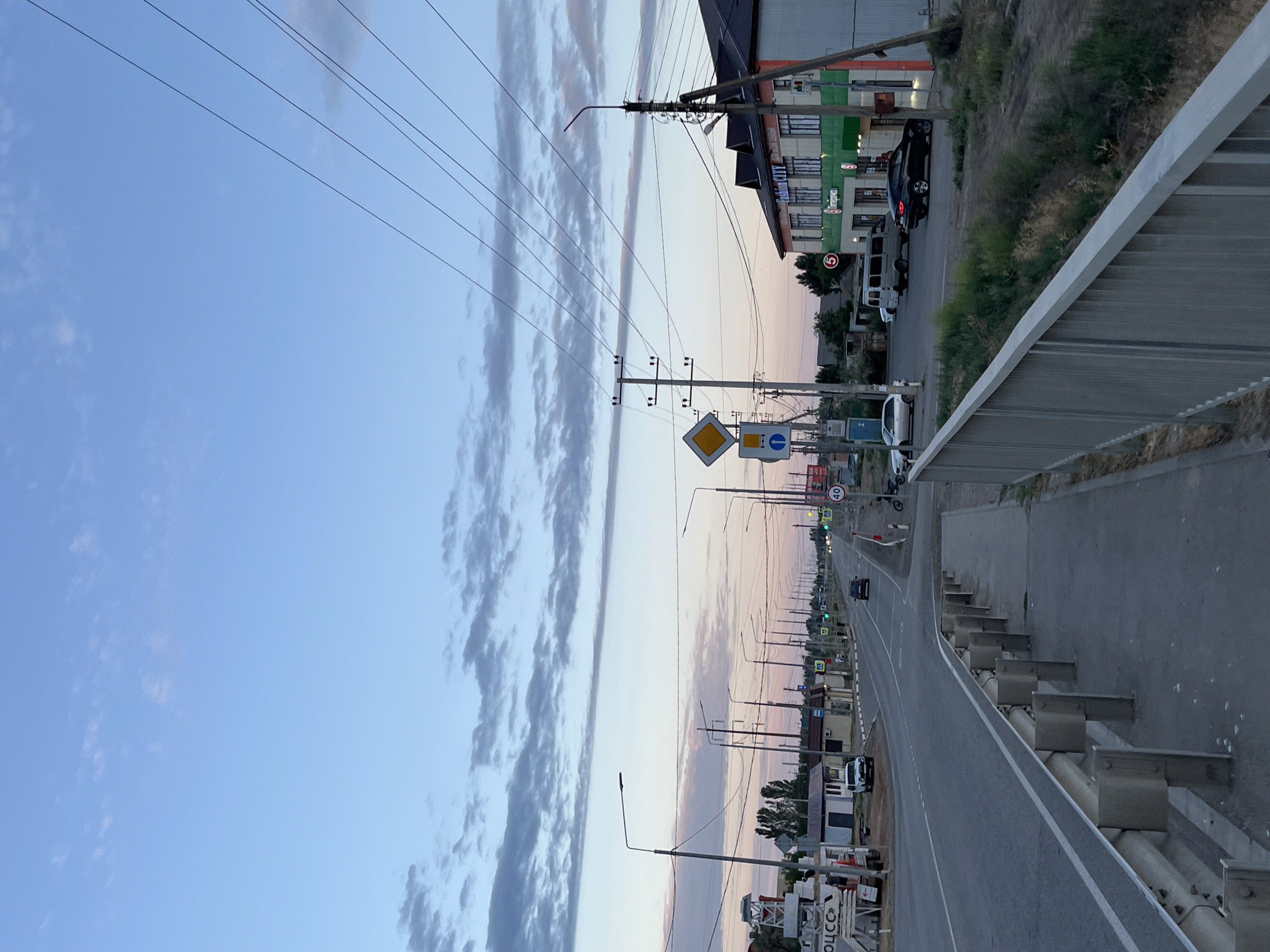 The image depicts a roadside scene at dusk in Krasnyye Barrikady, Russia. A two-lane asphalt road stretches into the distance, lined with various commercial buildings and streetlights. A pedestrian walkway with a metal railing is visible in the foreground. Several vehicles, including cars and vans, are parked or driving on the road. Overhead power lines crisscross the sky, which is tinged with soft hues of blue and pink from the setting sun. Traffic signs, including speed limit and directional signs, are present along the road. The overall environment suggests a typical peri-urban setting during the evening transition.