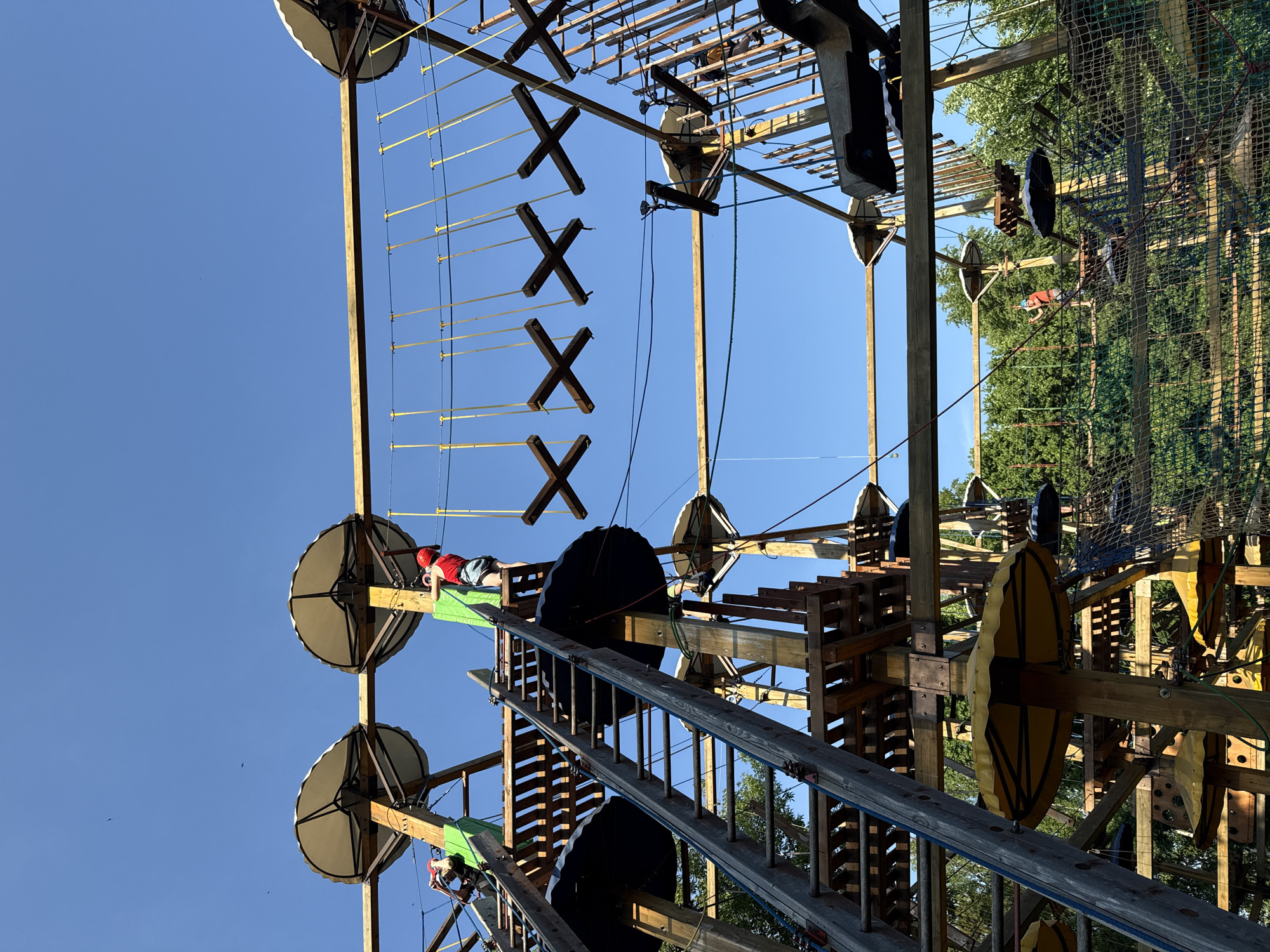 The image shows a treetop adventure park, possibly located in Izhevsk, Russia, although that cannot be confirmed. The park features multiple levels with a complex network of wooden platforms, ladders, ropes, and obstacles. Two individuals are visible navigating the course, wearing helmets and harnesses for safety. The park structure is built amidst trees. The sky is clear and blue.