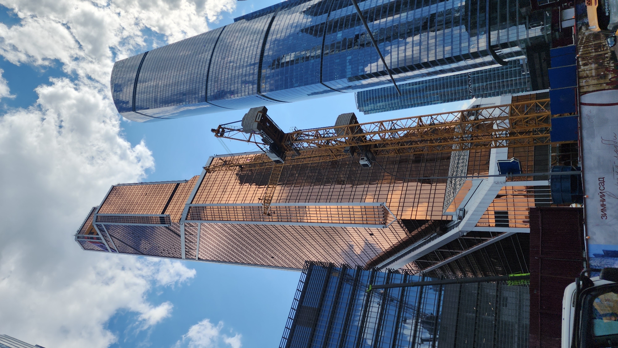 This is a low-angle, wide shot of a construction site with several modern skyscrapers under a cloudy blue sky. In the foreground, a yellow construction crane is positioned between two tall buildings. The building on the left is a skyscraper with a copper-colored glass facade, and it appears to be under construction, with scaffolding and construction elements visible. To the right of this building is a very tall, cylindrical skyscraper with a dark blue glass facade. Further in the background, other skyscrapers are visible, with some being taller than the foreground buildings. The sky has scattered white clouds. In the bottom foreground, a dark metal fence and a portion of a white vehicle are visible, along with a construction banner displaying Russian text. The overall impression is of a dense urban environment with ongoing development.