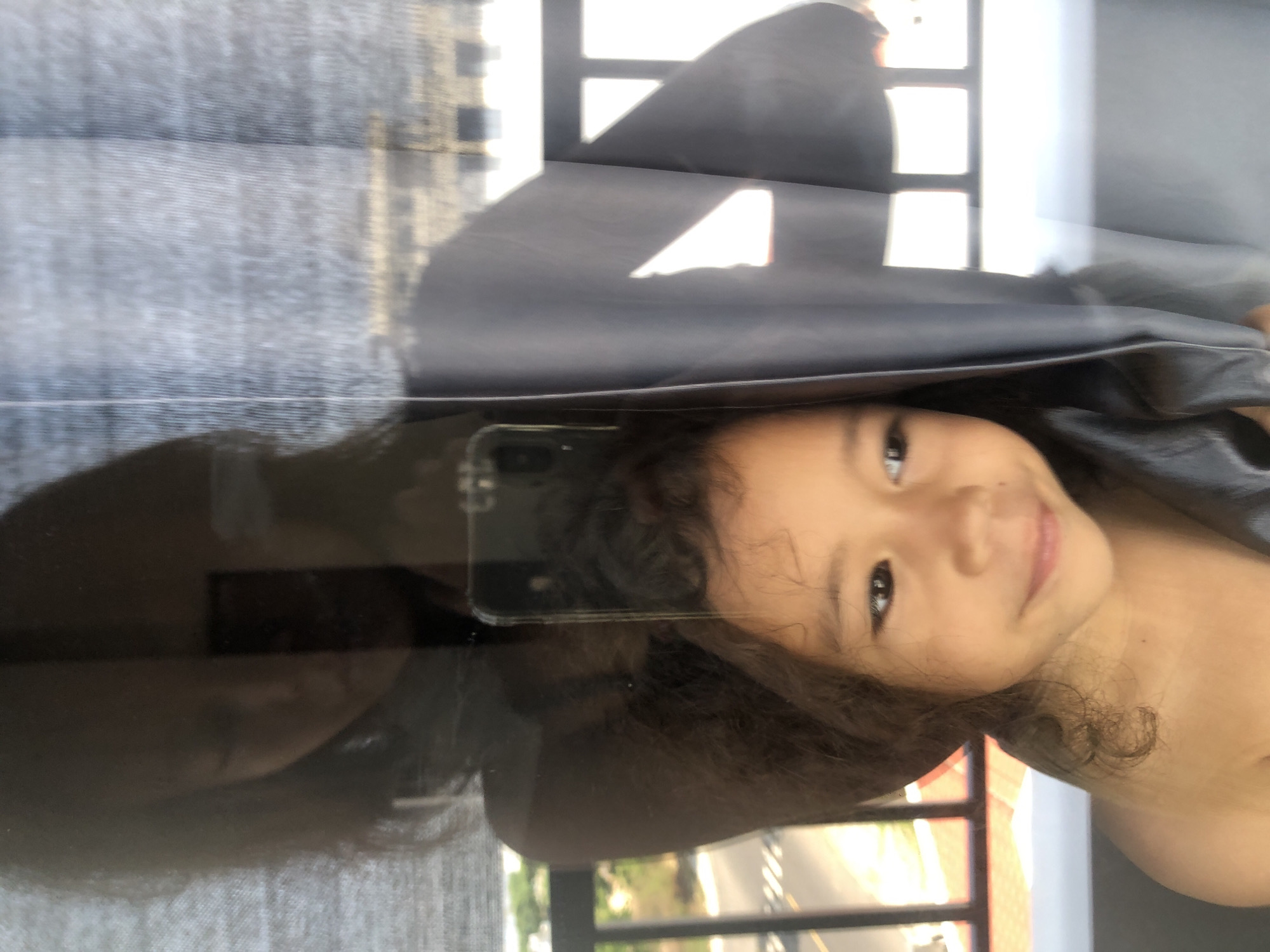 The image, oriented sideways, captures a young child's smiling face reflected on a glass surface, offering a superimposed view of interior and exterior elements. The primary subject is a child, likely a girl, with dark, curly hair, looking directly at the camera with a happy, slightly mischievous smile. Her face and part of her bare shoulder are visible, suggesting light attire.
Reflected on the glass near the child's head is a smartphone, indicating the photo might be a selfie or a capture of the reflection itself. To the child's left and above, a dark, possibly black or grey, fabric object with folds is visible. On the far left, a textured grey curtain or blind is reflected, partially obscuring the background.
Through and beyond the reflections, bright daylight illuminates the scene, streaming through distinct rectangular window frames. The outdoor view reveals an urban or suburban setting, with a road featuring white markings visible in the lower section, consistent with a city like Đà Nẵng, Vietnam. Distant buildings or structures can also be faintly discerned. The strong light suggests it is daytime. The image evokes a moment of playful interaction and happiness, blending indoor presence with an outward gaze. No visible text is present.