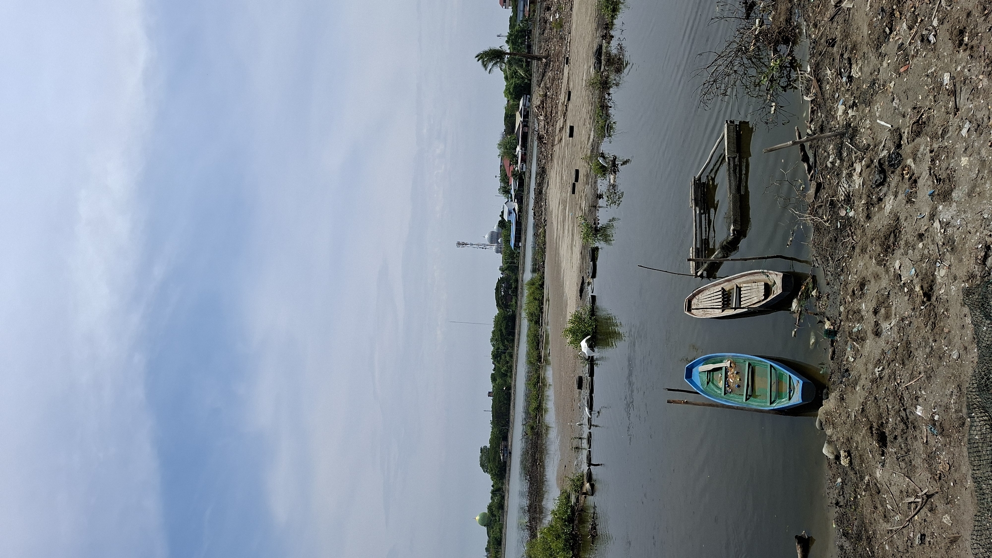 The image depicts a waterfront scene in Lhokseumawe, Indonesia. In the foreground, two small boats are moored in shallow water near a muddy bank littered with debris. One boat is blue and green, while the other is wooden. Further into the water, a submerged wooden structure is visible. The middle ground shows a muddy expanse with scattered vegetation, and several birds, including what appear to be egrets, are present. Beyond this area, a body of water separates the foreground from a distant shoreline. The distant shoreline is lined with trees and various buildings, including what appears to be a mosque with a green dome on the left and another mosque with white domes and a tall communication tower on the right. The sky is overcast with light clouds. The overall scene suggests a coastal or estuarine environment with signs of human habitation and potentially some environmental impact from debris.