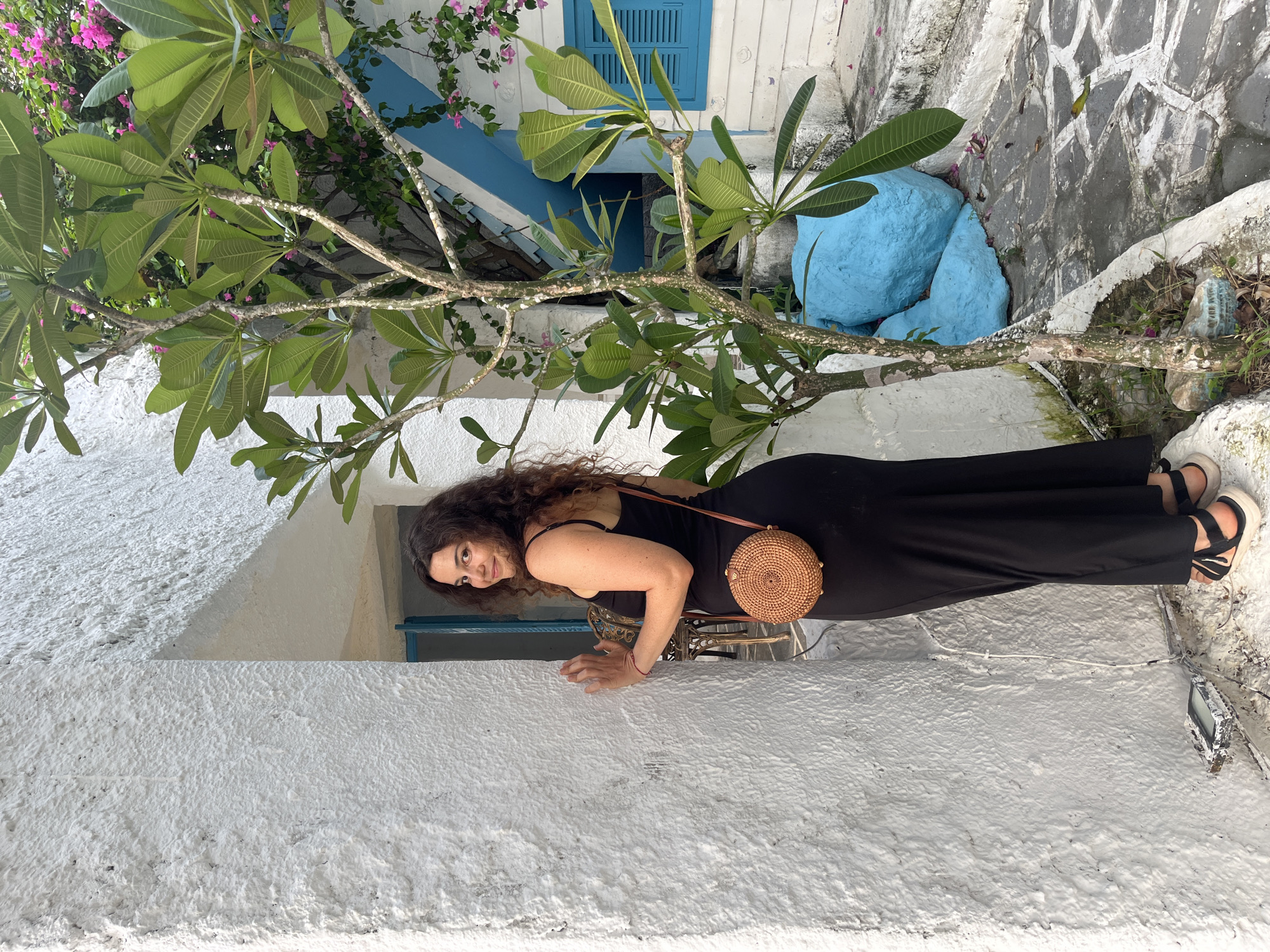 A woman with long, curly brown hair is leaning against a white textured wall, with her body angled towards the camera. She is wearing a black sleeveless dress and black sandals with thick white soles. A small, round, woven straw bag is slung across her body. Her right hand is resting on the wall in front of her, and her left hand is near her hip. She is looking directly at the camera with a slight smile.
Behind her, a tree with large green leaves and small pink flowers is visible. Further back, a building with white siding and blue trim and shutters is present. Two large, bright blue painted rocks are also part of the scene. The ground appears to be a mix of concrete and some dirt with dry vegetation. The image is taken in what appears to be an outdoor setting, possibly a courtyard or a street, with the style of the building and foliage suggesting a tropical or sub-tropical climate. The lighting suggests it is daytime. The overall mood is relaxed and casual. No visible text is present in the image.