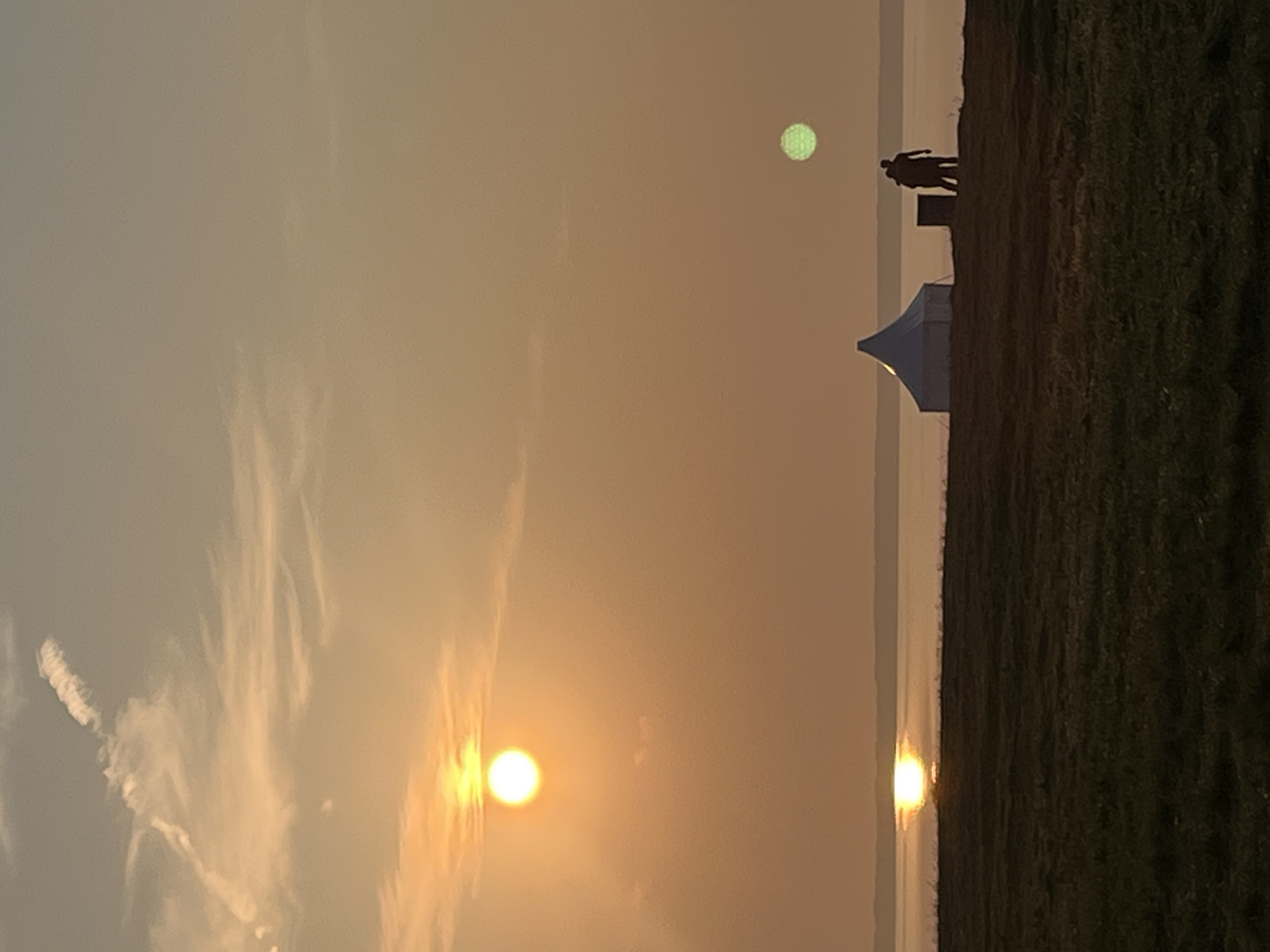The image depicts a tranquil sunset scene over a body of water. In the foreground, a grassy field stretches towards a gentle slope. On the slope, a white conical tent is visible, and to its right, two silhouetted figures stand close together, appearing to be a couple. Further out, the sun is setting, casting a warm orange glow across the sky and reflecting brightly on the water. A second, smaller green orb is present in the sky, possibly a lens flare or another atmospheric phenomenon. The horizon is marked by the distant line of the water meeting the sky. The location is identified as Surakovo, Russia.