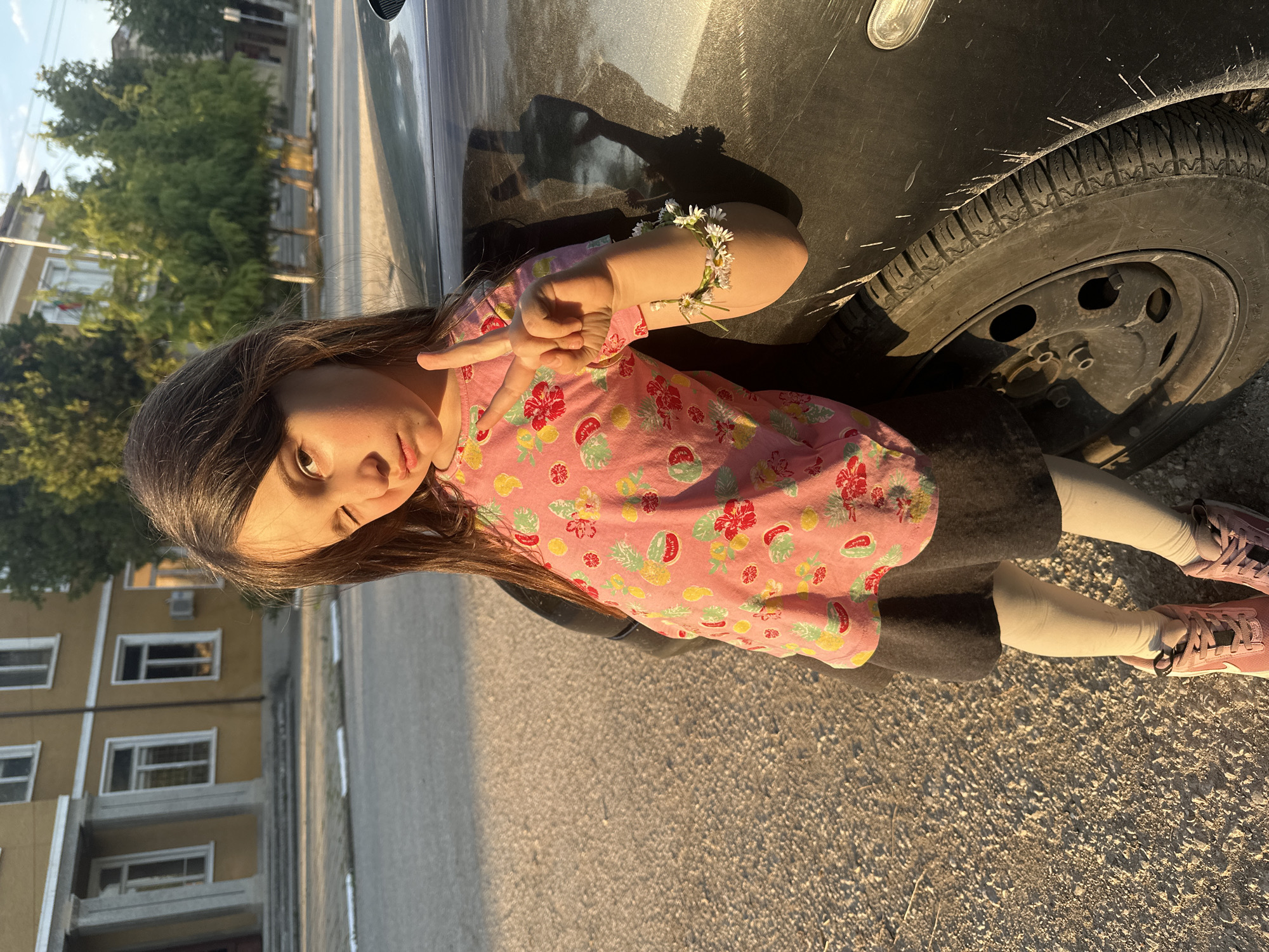 A young girl is leaning against a black car in Resen, Bulgaria. She is wearing a pink floral top, a grey skirt, white tights, and pink sneakers. She has a daisy bracelet on her arm and is holding up a peace sign with her fingers. The car tire is visible next to her. In the background, there is a building. The photo appears to be taken on a sunny day.
